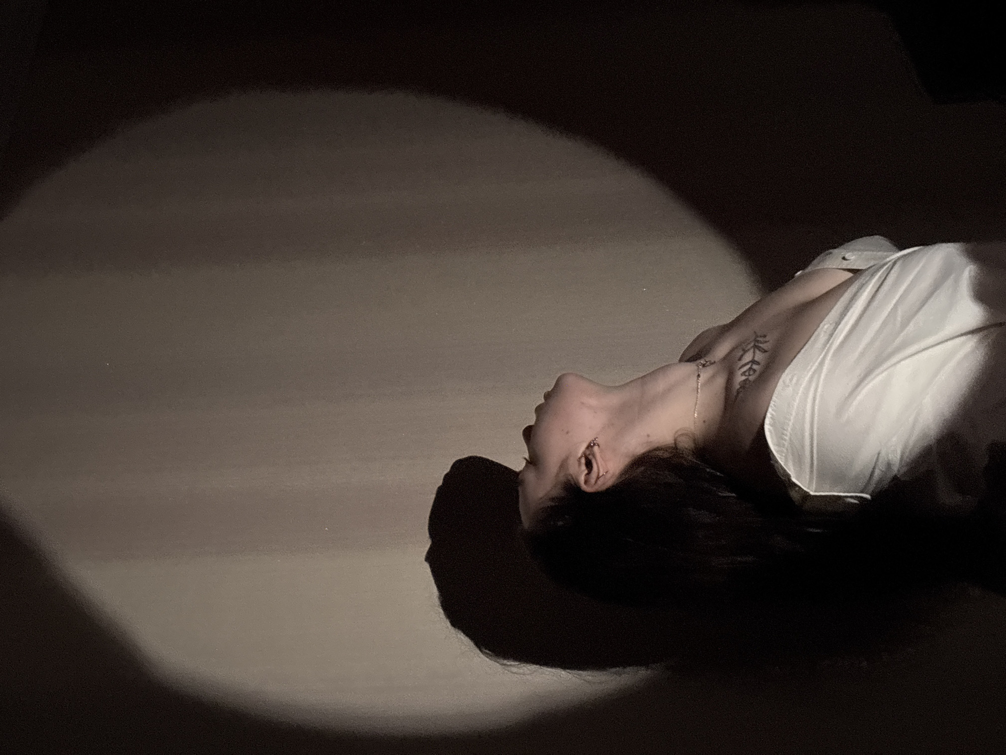 The image depicts a person lying down in a dimly lit room. A spotlight shines on the person, illuminating the face, neck, and upper chest. The person appears to be wearing a white shirt, and there is a tattoo visible on the neck. The background is dark, and the only other visible detail is the floor. There is no indication that the photo was taken in Perm, Russia.