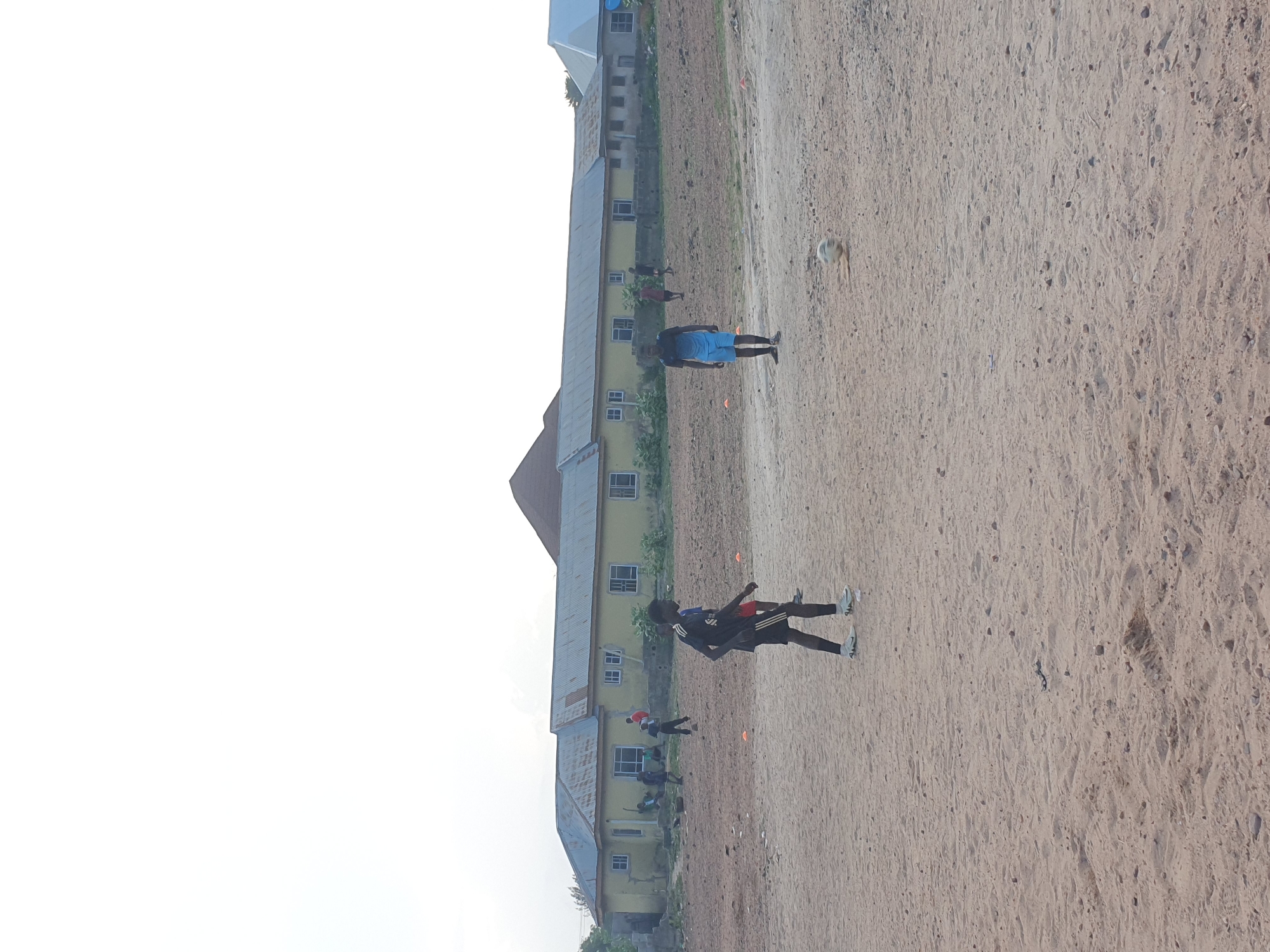 The image shows a dirt field with a yellow building in the background. Several people are present. One man is wearing a blue shirt and shorts, while another individual is wearing a black athletic outfit. Some people are running, while others appear to be walking. Orange cones are scattered across the field. The location is Lokoja, Nigeria.