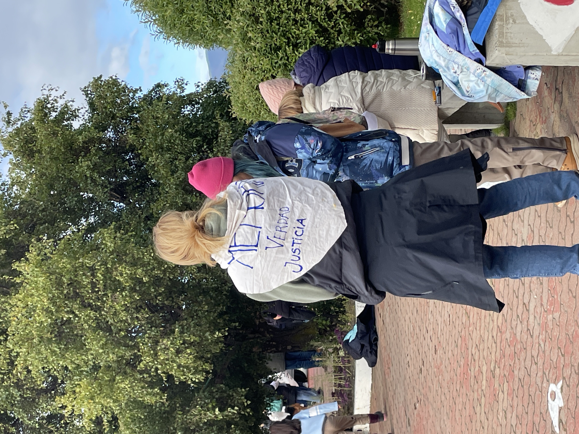 The image depicts an outdoor gathering in Ushuaia, Argentina. The central figure, viewed from behind, is a woman with blonde hair styled in a bun, wearing a light green hooded sweatshirt, a black sleeveless puffer vest, a long black skirt-like coat, and blue jeans. A white fabric item is affixed to her back, inscribed in blue with the text "MEMORIA VERDAD JUSTICIA." She stands on a reddish-brown paver pathway with an interlocking pattern.
To her right, a person wearing a bright pink knit beanie and a blue patterned winter jacket is partially visible, with blue hair extending from beneath the beanie. Further right, another individual wears a light pink knit beanie and a cream-colored quilted jacket, standing near a low concrete structure. On this structure, a silver thermal bottle with a red cap, a black mate cup with a bombilla, a lighter, and a folded blue and white patterned jacket are present.
In the mid-ground to the left, a child wearing a light blue and white striped shirt and light brown pants walks away from the viewer. Other indistinct figures are visible in the background beneath large green trees. The environment includes dense green foliage from trees and bushes. The sky is partially cloudy with patches of blue, and distant hills are visible on the horizon. A black item of clothing or bag lies on the pavers to the left of the central figure.