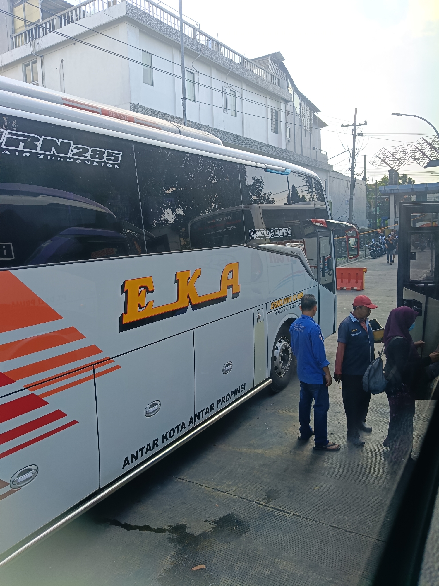 The image shows a white bus with orange and red stripes, labeled "EKA" in large yellow letters. The bus also has text that reads "RN 285 AIR SUSPENSION" and "ANTAR KOTA ANTAR PROPINSI". The scene appears to be a bus station or terminal in Kedungrejo, Indonesia. There are three people standing next to the bus: a man in a blue shirt and jeans, a man in a navy shirt with red trim and a red cap, and a woman wearing a purple hijab. The man in the red cap is holding a yellow ticket dispenser. The bus is parked next to a building, and there are other vehicles, including motorcycles, visible in the background. The ground is paved with concrete.