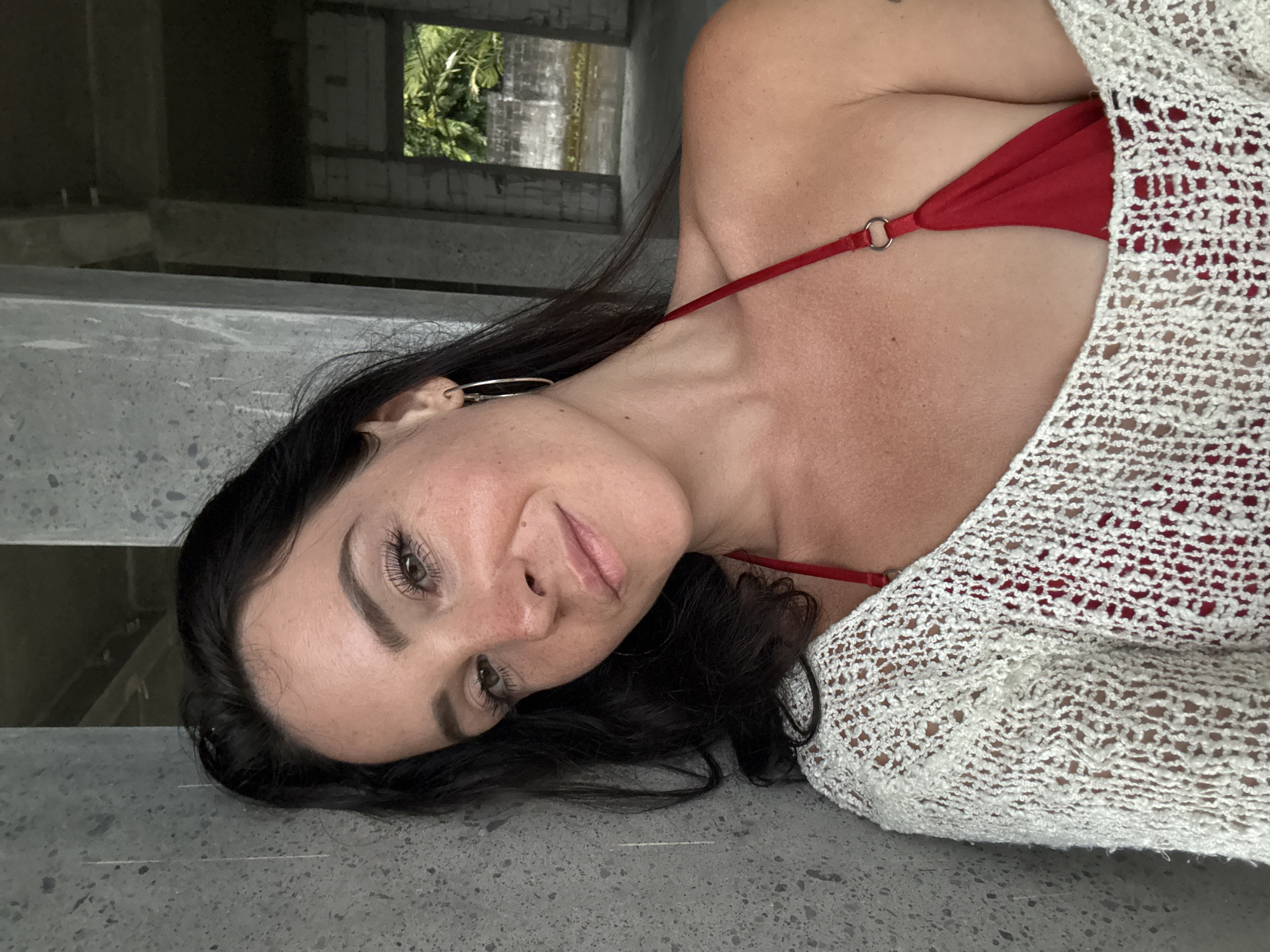 A woman with dark hair and light skin poses for a selfie. She is wearing a red bikini top and a white, open-knit sweater. She is leaning against a concrete wall, with an unfinished building and foliage visible in the background. The scene appears to be outdoors in a warm climate, possibly Bebandem, Indonesia.