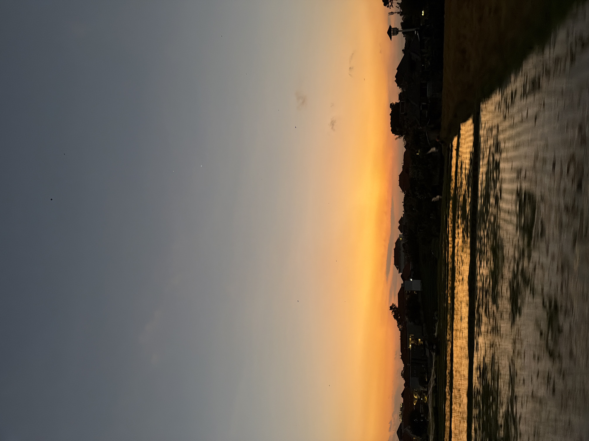 The image depicts a rural-to-semi-urban landscape at dusk in North Kuta, Indonesia. The sky transitions from a deep grey-blue at the zenith to a vibrant orange and yellow hue along the horizon, characteristic of a sunset. Sparse, indistinct clouds are present, primarily in the middle sky. Several small, dark specks, possibly birds or kites, are scattered across the sky, along with a few bright, pinpoint lights, likely stars or planets. Below the horizon, a silhouette of buildings with traditional Balinese architectural elements and surrounding trees is visible. Some interior lights illuminate portions of these structures. The foreground is dominated by flooded rice paddies, their surface reflecting the warm colors of the sunset in horizontal streaks, indicating either slight water movement or motion blur from the photographic capture. To the far right, a section of dry land borders the paddies. No discernible individuals or specific human activities are present within the frame.