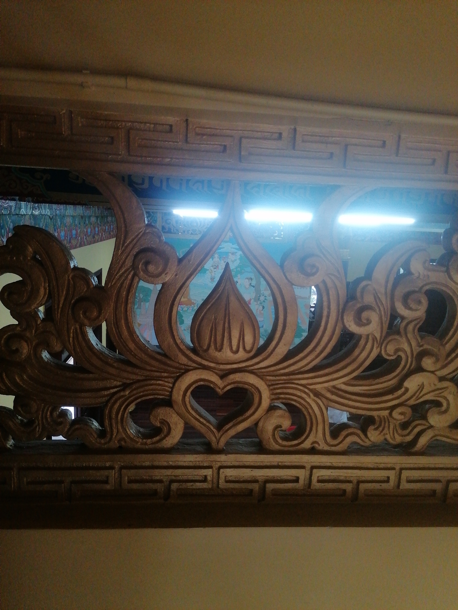 The image shows a close-up of ornate wooden carvings, likely part of a decorative architectural element, such as a screen or a railing. The carving features intricate patterns, including swirling motifs, stylized floral elements, and a central heart shape. Behind the carving, a colorful mural or tapestry is visible, depicting figures and possibly a landscape. Two bright fluorescent lights are illuminating the scene from above, creating a strong glare. The overall impression is one of detailed craftsmanship and cultural artistry. The specific location in Da Lat, Vietnam, is not discernible from the image itself.