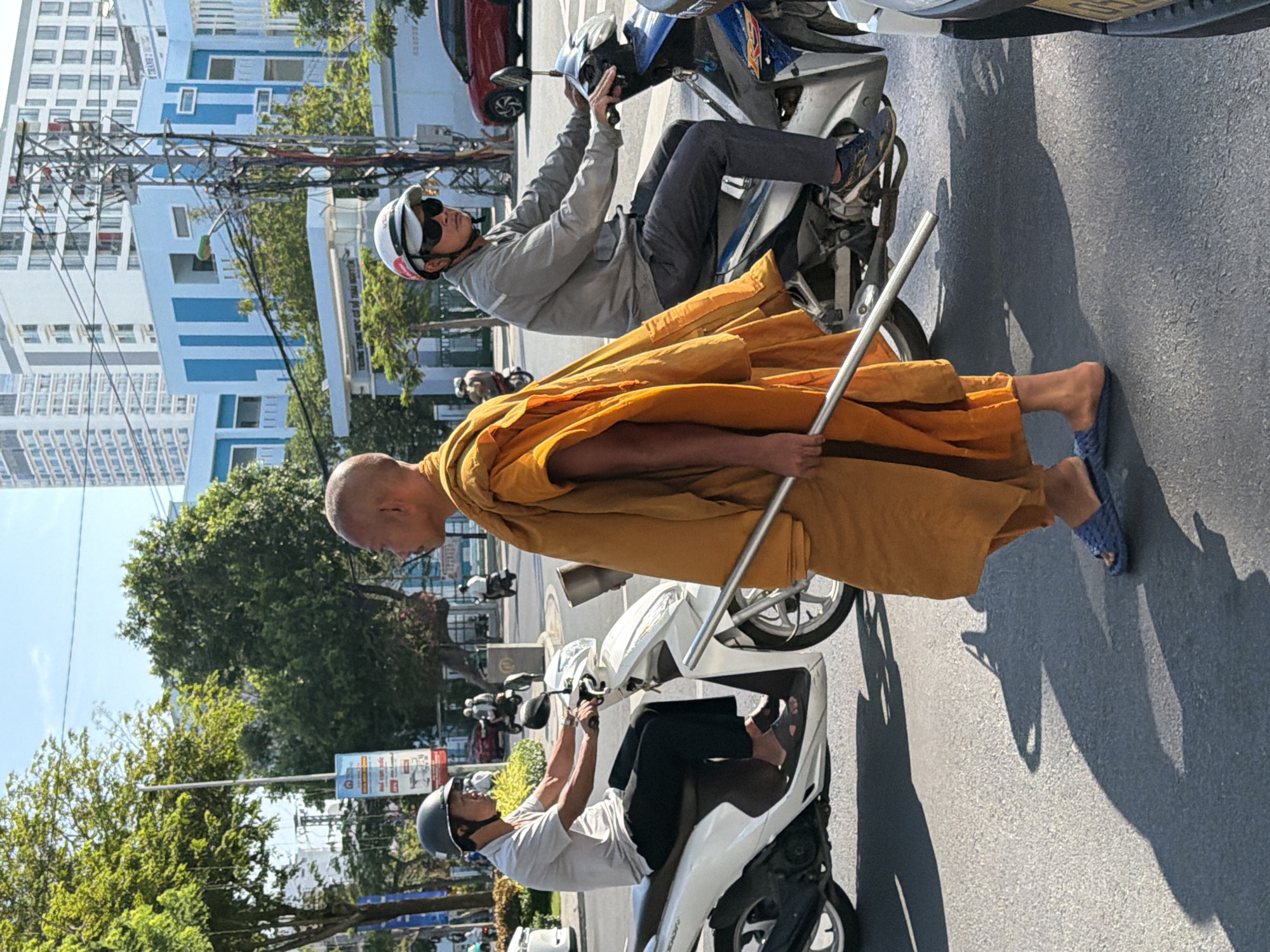 In Nha Trang, Vietnam, on a sunny day, a Buddhist monk in saffron robes walks across a street, holding a metal staff. He is wearing blue slippers. To his left, a man wearing a grey shirt, black pants, a helmet, and sunglasses rides a white scooter. To his right, another man, also wearing a helmet and sunglasses, rides a blue and silver scooter. In the background, modern buildings, trees, and other vehicles are visible. The scene captures a moment of daily life in the city, with a blend of religious and secular elements.