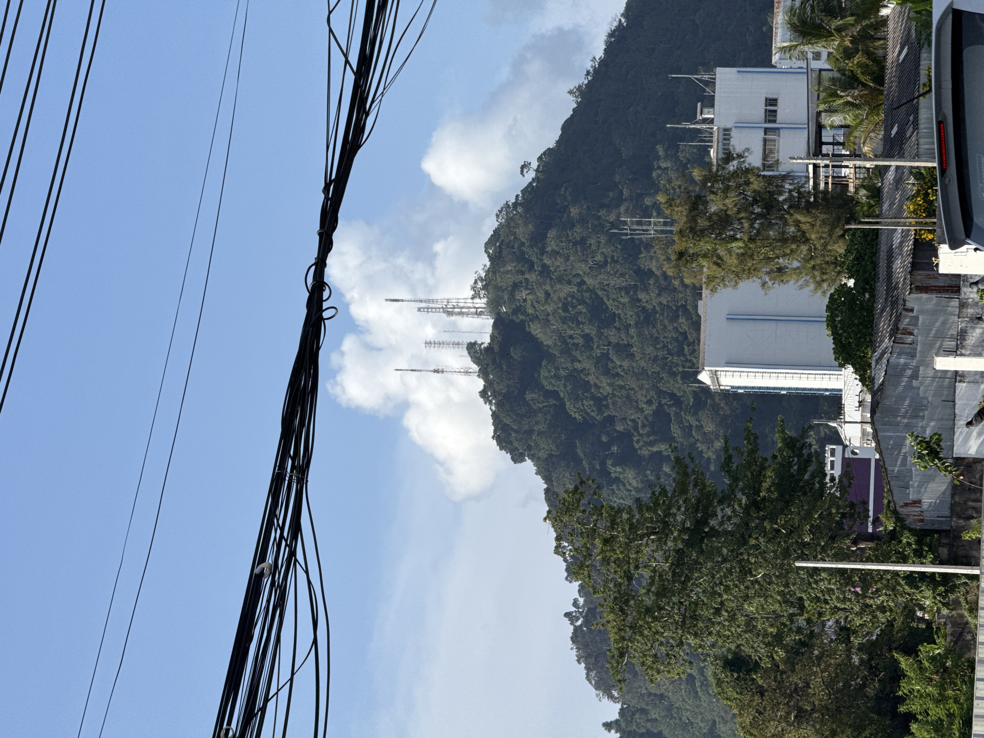 A wide shot shows a lush, tree-covered mountain in Phuket City Municipality, Thailand, topped with several communication towers. Power lines crisscross the foreground against a blue sky with scattered clouds. In the mid-ground, a white, multi-story building with blue accents is visible, along with corrugated metal roofs of smaller structures. The overall scene suggests an urban environment encroaching on a natural landscape.