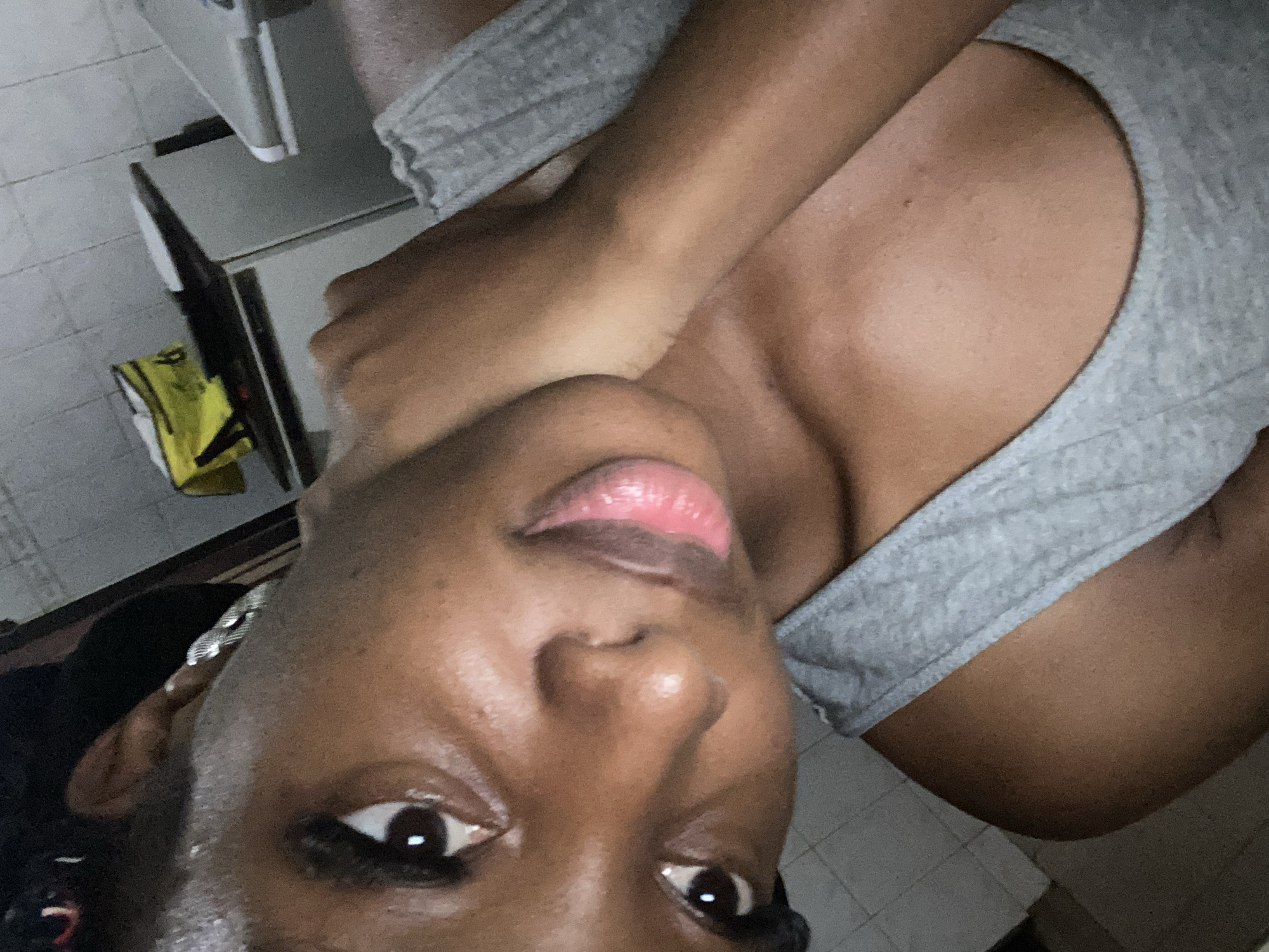 The image shows a close-up, angled selfie of a young Black woman wearing a gray tank top. Her head is tilted, and her left hand is touching her neck. She has makeup on her eyes and lips, which are a bright pink color. The background appears to be a bathroom with tiled walls and what looks like a kitchen counter with a yellow bag on it. The lighting is dim, creating a slightly hazy effect. There is no discernible location in the city of Jos, Nigeria, mentioned or depicted in the image.