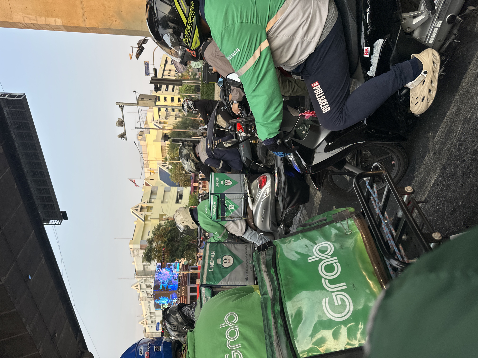 The image shows a row of parked motorcycles in what appears to be Bangkok, Thailand. Delivery riders, identified by their "Grab" and "LINEMAN" branded backpacks and apparel, are sitting on their bikes. The riders are wearing helmets and jackets. The bikes are parked alongside a curb, with buildings and a bridge structure visible in the background. The delivery riders are likely waiting for assignments or orders. The presence of the police department can be observed.