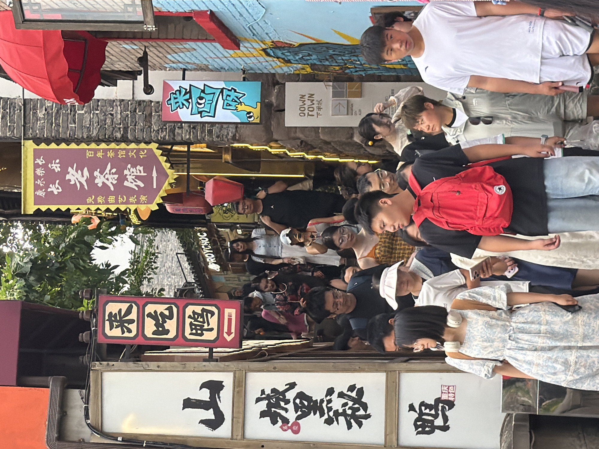 The image depicts a busy street scene in Yuzhong District, China. A crowd of people are walking up a narrow, stone-paved alleyway lined with traditional Chinese architecture. Various signs with Chinese characters are visible, including one for "Roy Choc" and another with playful cartoon characters. The signs suggest a commercial area, possibly a market or a tourist street. The people in the image are dressed in casual clothing, indicating a daytime scene. Some are carrying bags or backpacks, and a few are looking at their phones. The atmosphere appears lively and crowded, typical of a popular urban spot. The buildings are multi-storied with dark tiled roofs and brick facades. The overall impression is one of a vibrant and bustling pedestrian street.
