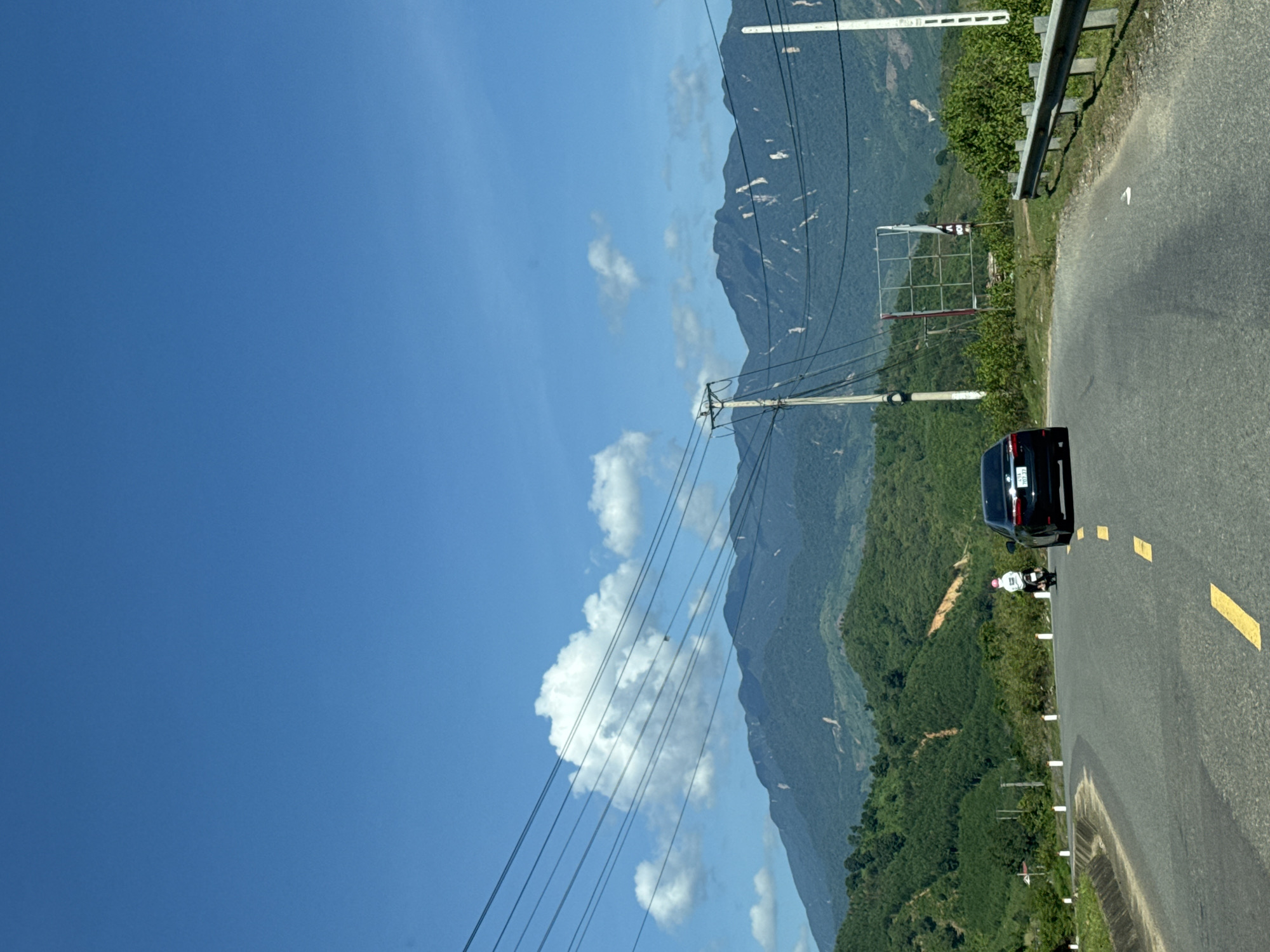 A black sedan with license plate 703.37 is traveling on a paved road. A person on a motorcycle, wearing a white shirt and pink helmet, is ahead of the car. The road is bordered by a metal guardrail on the right and grassy shoulders with white posts on the left. In the background, lush green hills rise up to meet a bright blue sky with scattered white clouds. Power lines stretch across the scene, supported by utility poles. A large, empty billboard structure is visible on the right side of the road. The overall scene depicts a scenic drive through a mountainous, rural area, likely in or near Nam Khánh Vĩnh, Vietnam, given the context of the request.