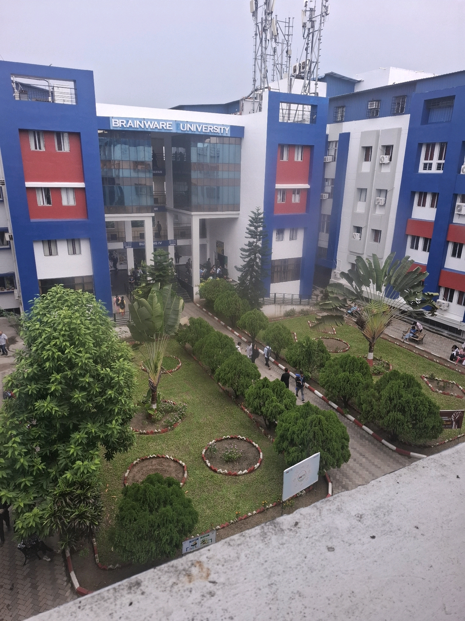 The image captures a view of the Brainware University campus in Barasat, India. The university building is modern, with a striking blue and red facade. Large glass windows dominate the central section of the building, reflecting the sky. In front of the building, a well-maintained garden features manicured bushes, a large palm-like tree, and a grassy lawn. Several people are visible walking on pathways and sitting on benches within the garden area. The campus appears to be bustling with activity, suggesting it is a normal day for students and staff. The overall impression is one of an educational institution set within a pleasant, green environment.