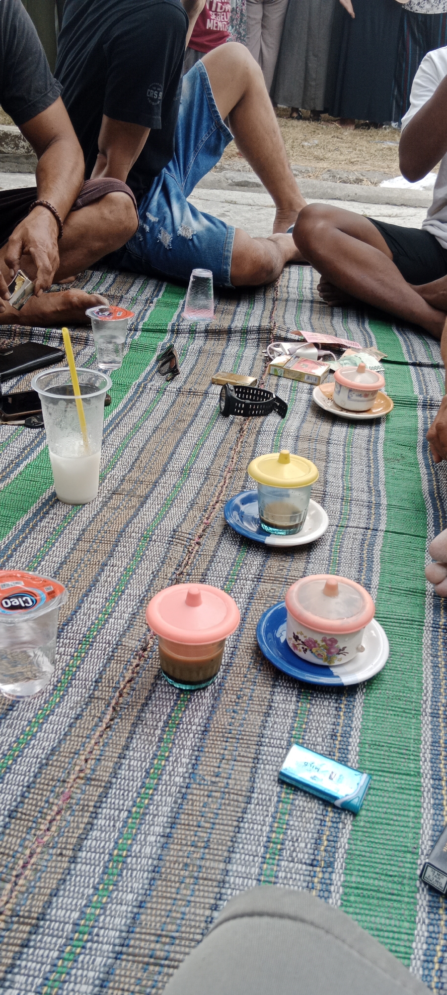 The image depicts a casual gathering of at least three individuals seated on a woven mat outdoors, likely in Gintungan, Indonesia. Two individuals are visible from the waist down, one wearing blue distressed denim shorts and the other in black shorts. A third person, partially visible, wears a white t-shirt. They are seated on a multi-colored woven mat with stripes of green, blue, and beige.

Scattered on the mat are various items. In the foreground, a clear plastic cup with a yellow straw contains a milky white beverage. To its left is a clear plastic cup with a red lid, and a partially visible brand name "CLEO" is visible on its side. Further to the right, there are two small cups with lids. One has a pink lid and contains a dark liquid, possibly coffee, and is placed on a blue and white saucer. Another similar cup with a pink lid also contains a dark liquid and sits on a floral-patterned saucer. Adjacent to this is a glass with a yellow lid containing a dark liquid, on a blue saucer.

Other objects on the mat include a pack of cigarettes, a lighter, and a black wristwatch. In the background, other individuals are partially visible, some wearing traditional Indonesian attire. The ground surrounding the mat appears to be dry earth or sand. The overall impression is a relaxed, informal social setting.