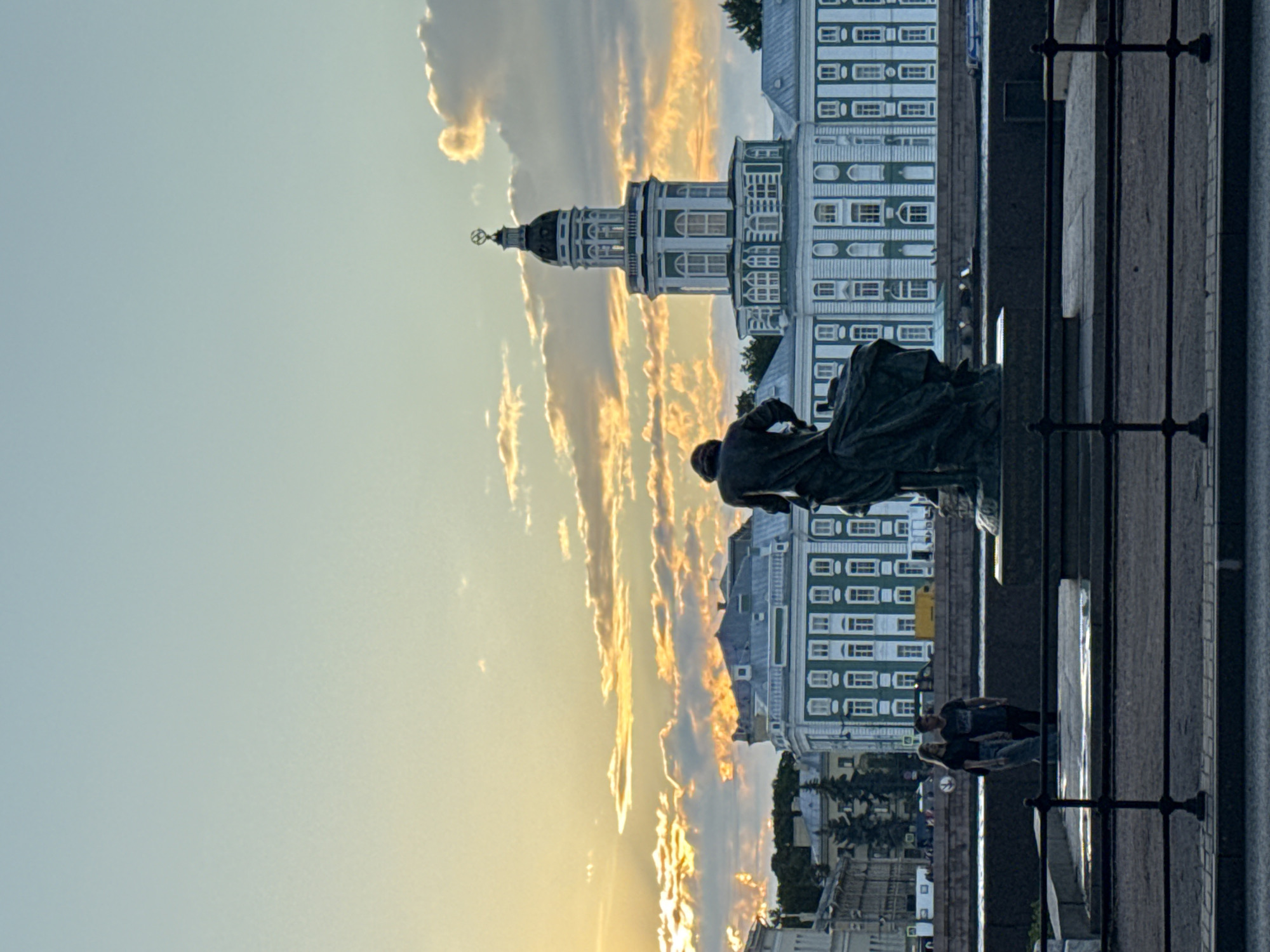 The image shows a wide shot of the Kunstkamera museum in Saint Petersburg, Russia, during sunset. The museum's distinctive green and white facade is visible, with its central tower topped by a globe and compass. In the foreground, a dark bronze statue of a man in flowing robes stands on a pedestal. A couple, a man and a woman, are standing on the embankment to the left of the statue, looking towards the river. The sky is filled with dramatic clouds illuminated by the setting sun, casting a warm glow over the scene. The river Neva flows in front of the museum, with a dark railing in the foreground. The overall atmosphere is serene and picturesque, capturing the grandeur of the city at dusk.