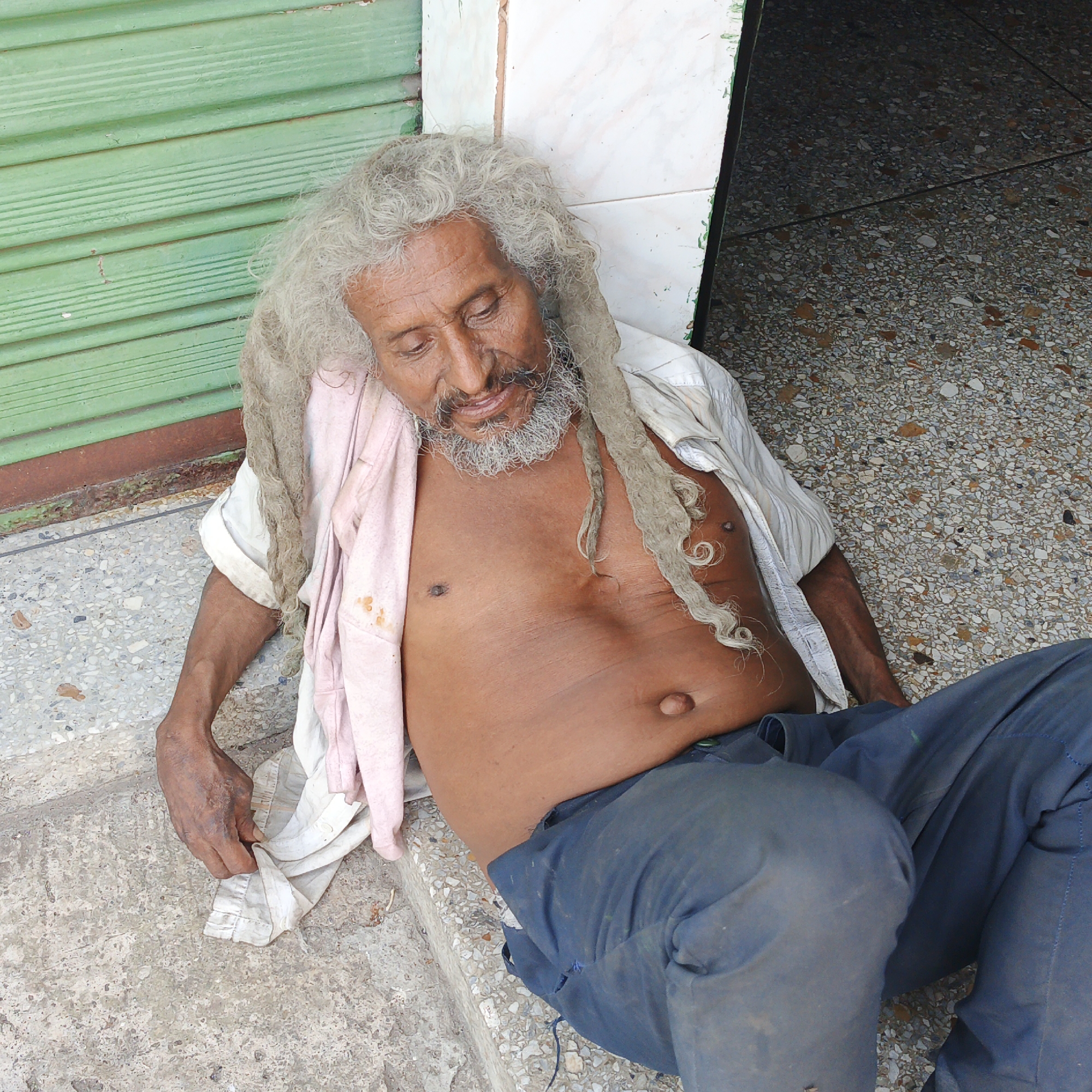 This image depicts an adult male in Tinaquillo, Venezuela, positioned on a speckled concrete surface and leaning against a building facade. The individual has a dark complexion, a heavily wrinkled face, and long, matted, light gray hair forming dreadlocks, accompanied by a matching gray beard and mustache. His eyes are downcast. He is shirtless, exposing his chest and abdomen, which features a protruding navel. A rumpled white long-sleeved shirt is partially draped over his left shoulder and arm, underneath a light pink fabric or garment that displays reddish-brown stains. He wears dark blue trousers. His left hand and forearm rest on the ground. The immediate background to the left consists of a green horizontally-slatted structure, consistent with a metal rolling shutter. To the right, a white tiled wall meets a dark recessed area or doorway, revealing a dark, multi-colored aggregate floor. The individual's posture suggests a state of rest or unconsciousness.
