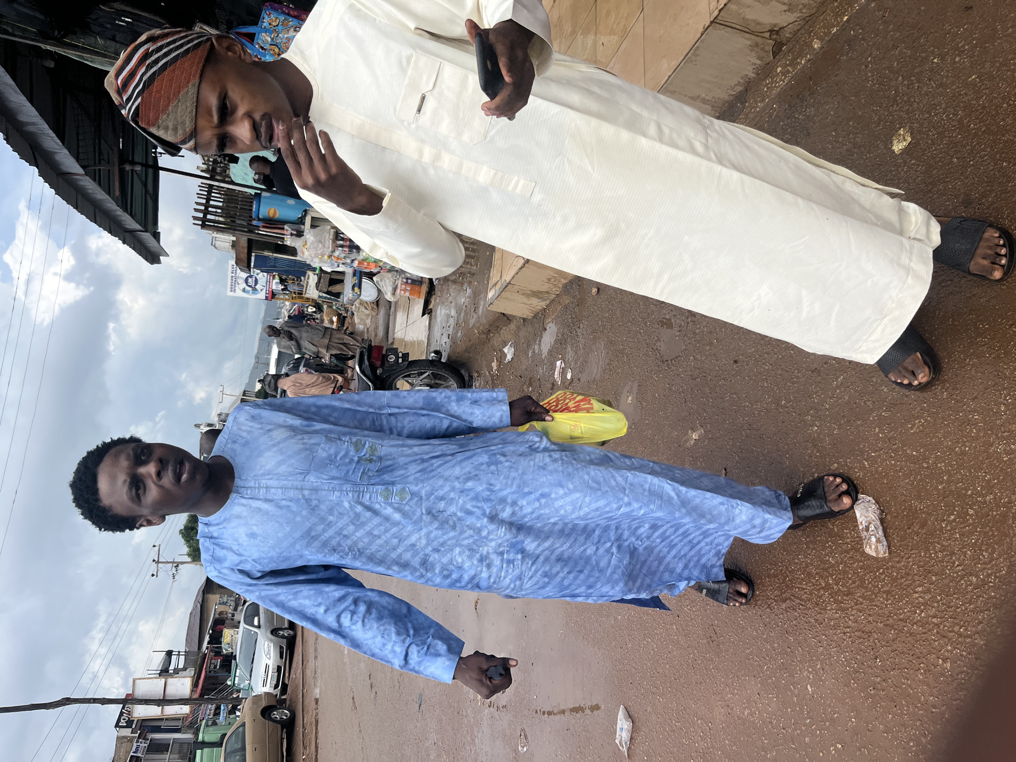 Two men are walking on a wet, reddish-brown street in Jos, Nigeria. The man on the left wears a long, light blue patterned robe and black sandals. He holds a small, dark object in his right hand and a yellow plastic bag in his left. He is looking to his left and speaking. The man on the right wears a cream-colored robe and black sandals. He has a patterned cap on his head. His right hand is near his mouth, and he is holding a dark mobile phone in his left hand. He is looking to his right. In the background, there are several shops with awnings and a few cars. The sky is overcast.
