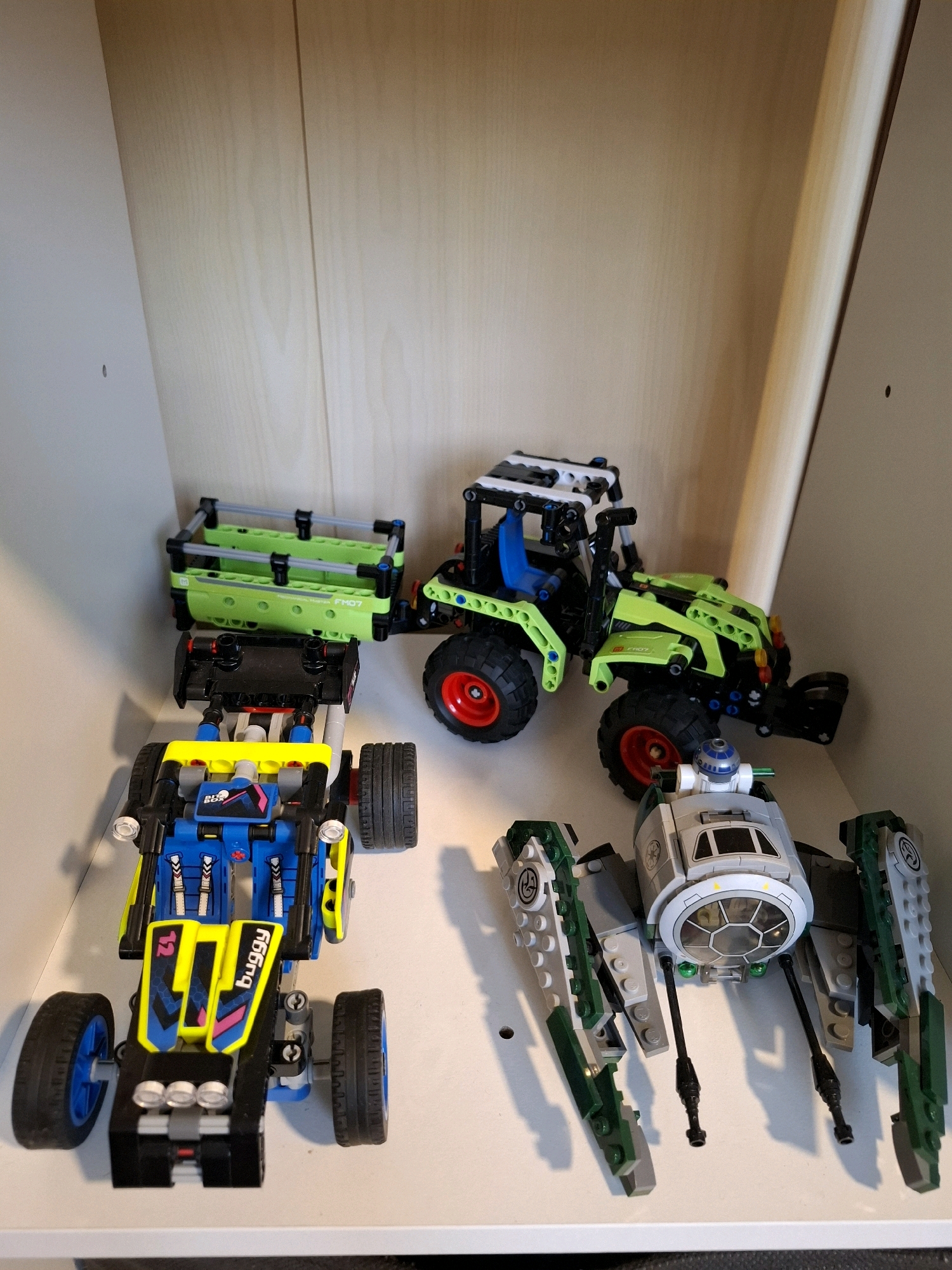 The image shows a collection of LEGO toys on a white shelf. In the foreground, a blue and yellow LEGO buggy is positioned. Behind it, a green LEGO tractor is visible. To the right of the tractor, a grey and green LEGO spaceship with a R2-D2 figure is partially shown. The background is a light-colored wooden wall, and the scene appears to be indoors, possibly in a home. There is no indication of the city of Tyumen, Russia, or any specific urban context.