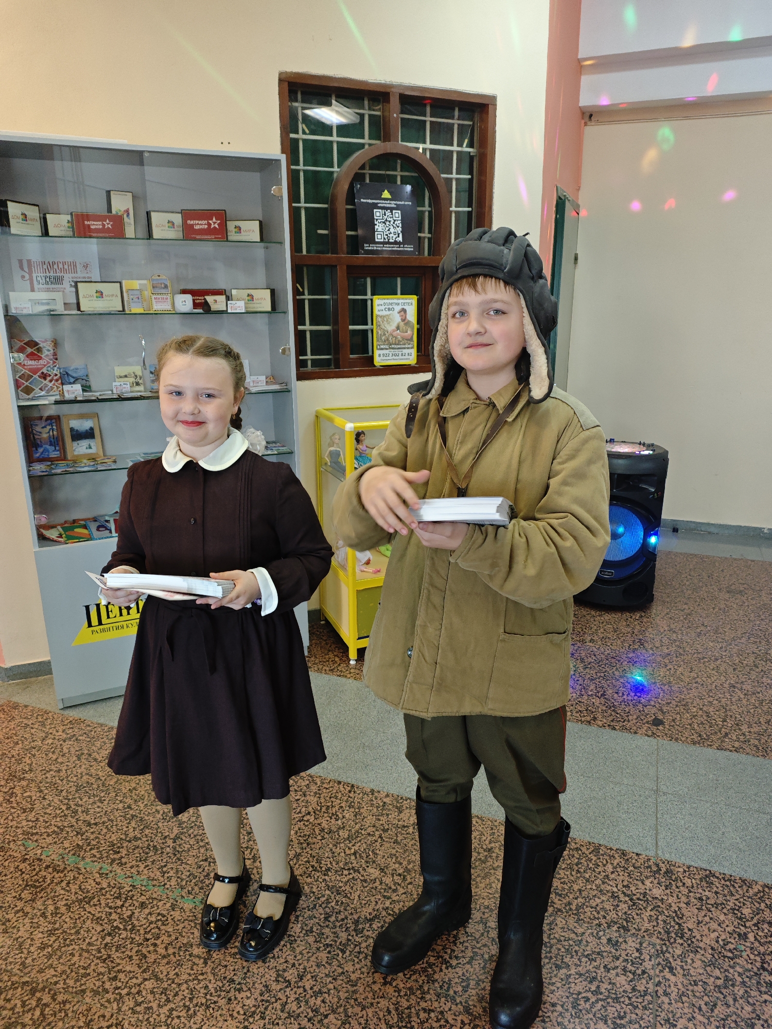 Two children are standing in what appears to be a building interior, possibly a cultural center or school based on the decor. The girl on the left wears a brown dress with a white collar and black shoes, and holds a stack of white papers. To the right stands a boy dressed in a military-style outfit, including a tan jacket, green pants, black boots, and a pilot's hat with ear flaps. He is also holding a stack of papers.
Behind them is a display case filled with various items, including signs with the text "ПАТРИОТ" and "МИРА". A poster and a QR code are visible on a window behind the boy. There is also a yellow storage unit with dolls on display and a blue-lit speaker behind the boy.
The floor has a speckled brown and gray pattern.
Based on the signage in the image, particularly the Cyrillic text, the location is Markovskii, Russia. The text "Для ОПЛЕТКИ СЕТЕЙ для СВО" on the poster indicates support or fundraising for special military operations. The presence of the QR code suggests digital information access.