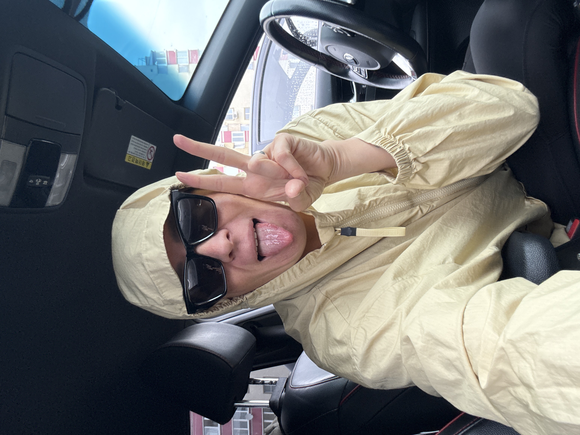 A person sits in the driver's seat of a car in Chaikovsky, Russia, sticking out their tongue and making a peace sign with their right hand. The person is wearing a light yellow hooded jacket and black sunglasses. The interior of the car is visible, including the steering wheel, dashboard, and a part of the passenger seat. Outside the car, buildings and a cloudy sky can be seen.