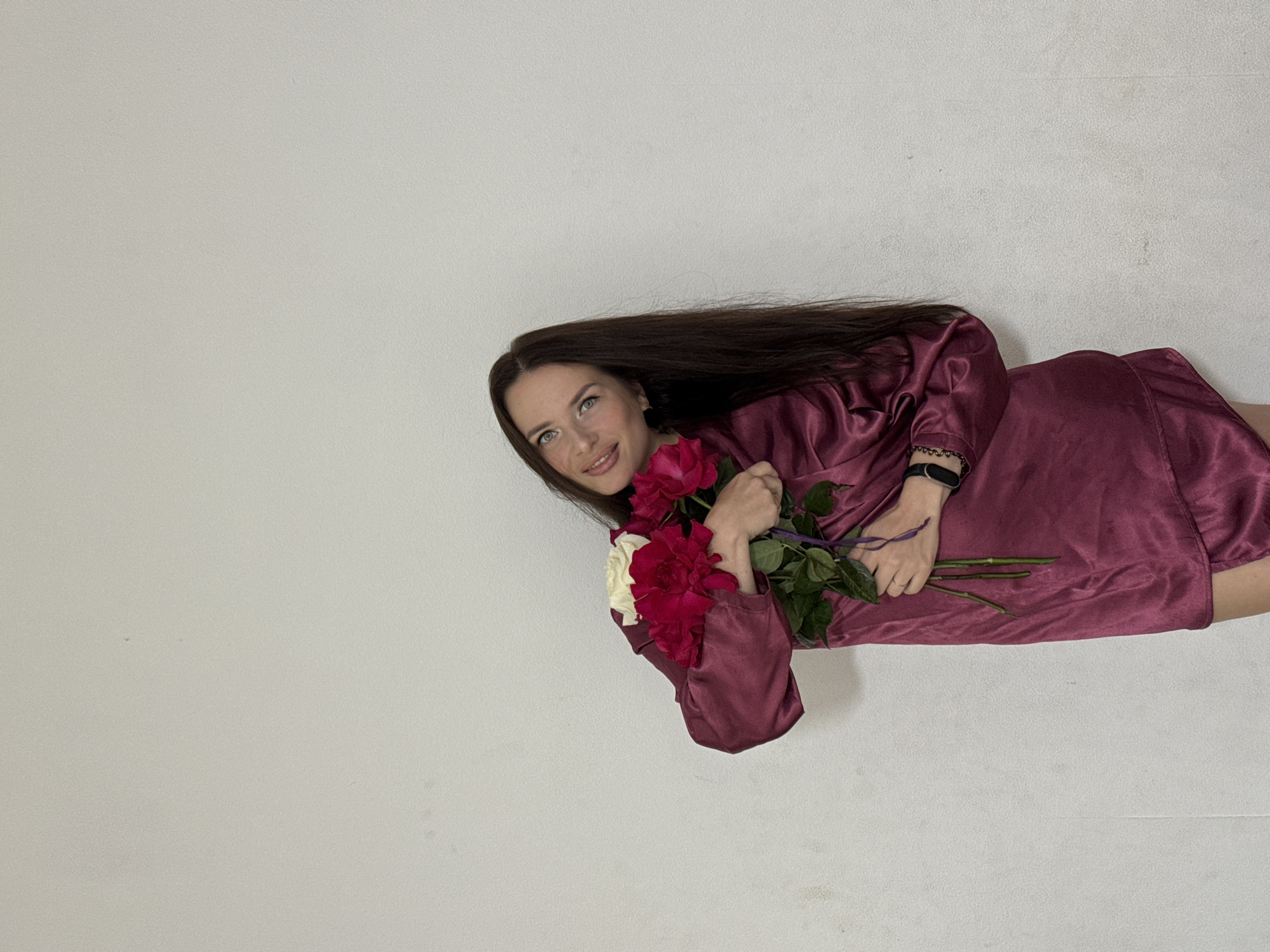 A young woman with long, dark hair and blue eyes is standing against a plain, off-white wall. She is holding a bouquet of red and white roses. She is wearing a dark pink, silky pajama set. The woman is smiling and looking towards the camera. The background is a simple, textured wall. The location is identified as Rusalevka, Russia.