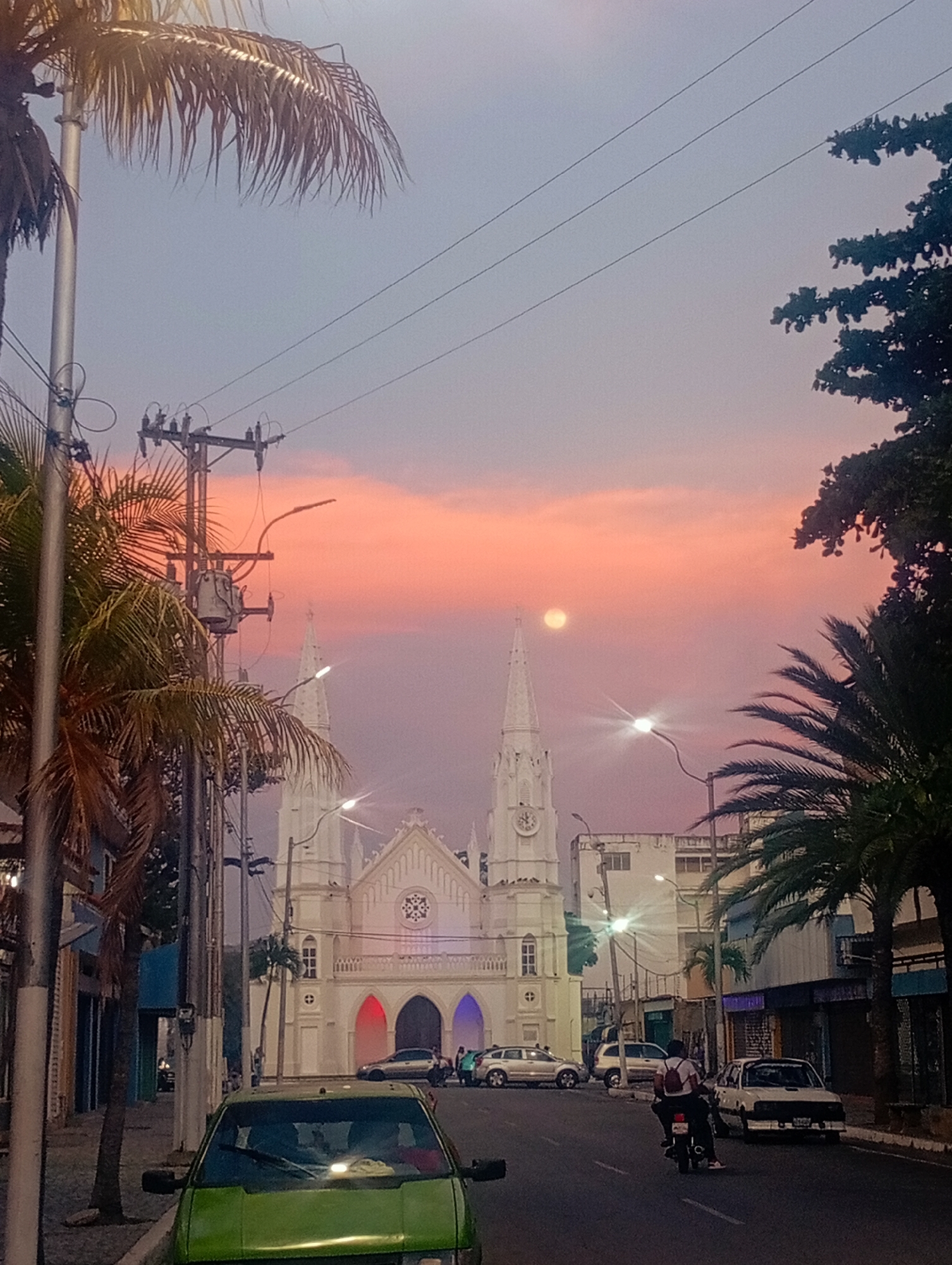 The image presents a striking twilight scene on a street in Juan Griego, Venezuela, looking towards a prominent white church. The sky is dominated by vibrant orange and pink clouds, indicating either sunset or early dawn, with a faint moon visible amidst the colorful expanse. Streetlights are illuminated, casting bright glares and signifying the transition to night.
At the heart of the composition is a grand, white church, distinguished by its two tall, ornate spires, each featuring a clock face. The church's facade showcases arched entrances, with some arches illuminated in hues of red, blue, and purple, adding a festive glow. A circular decorative window is positioned above the central entrance.
The street is moderately active with vehicles. A bright green car is parked prominently in the foreground on the left. Further down the road, near the church, a silver car and a white car are parked. A person on a motorcycle, wearing a backpack, is seen riding away from the viewer on the right side of the street. A few other indistinct figures are visible near the church entrance and on the sidewalk.
Palm trees, with their fronds silhouetted against the sky, frame both sides of the scene, contributing to a tropical atmosphere. Utility poles, transformers, and a network of overhead electrical wires are prominent on the left and across the sky, characteristic of an urban environment. Various buildings, consistent with a town setting, line the street. The overall scene captures the serene yet active essence of a town at a beautiful transitional hour of the day. No legible text is visible within the image.