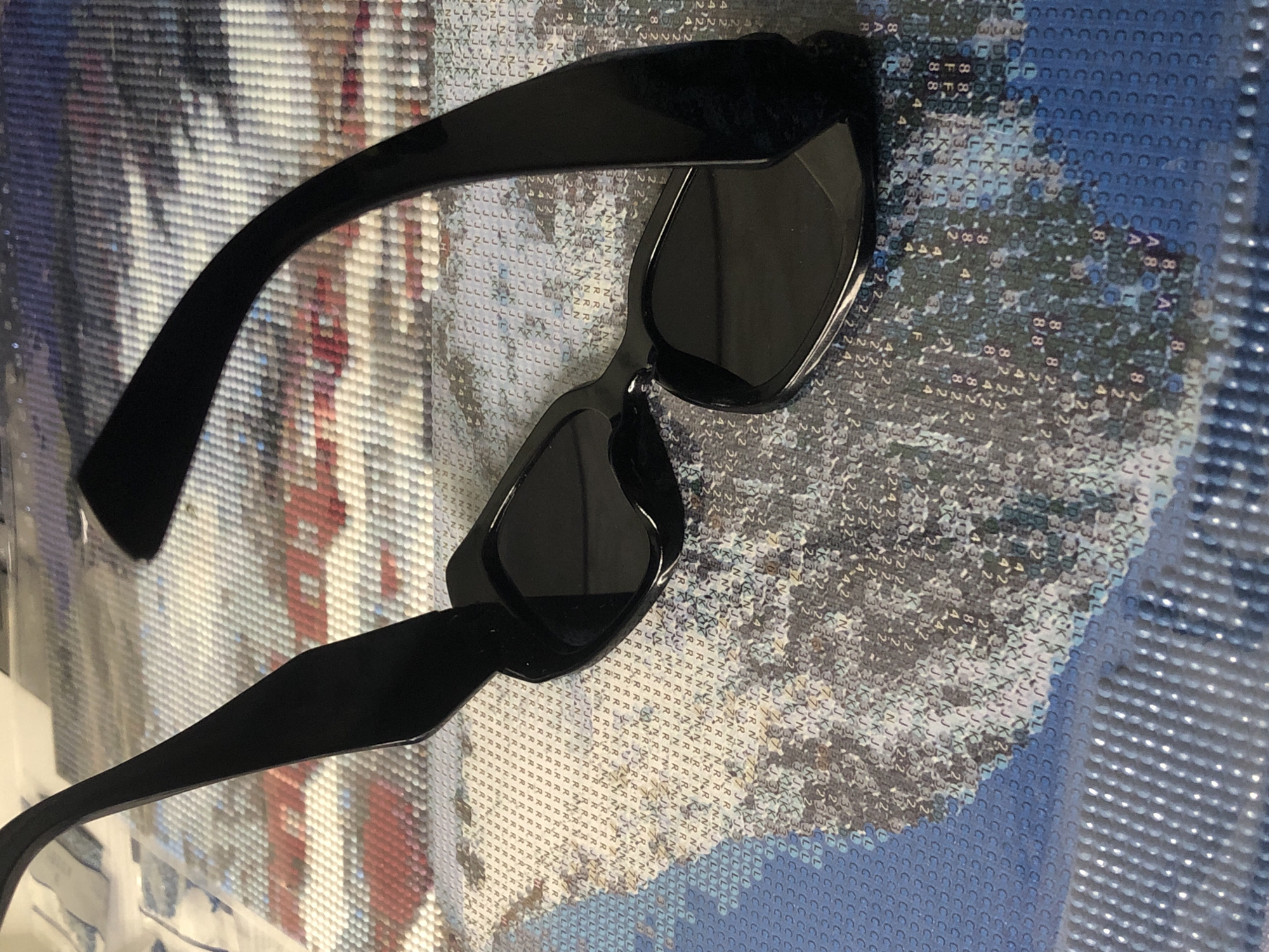 The image shows a pair of black sunglasses resting on a surface with a mosaic-like pattern of small shapes. The mosaic includes colors such as blue, white, grey, red, and dark shades. The sunglasses are angled to the right, with one arm extending upwards and the other downwards. The sunglasses appear to be modern in style. The environment has no clear indicators to identify the location in Noviy, Russia, and the absence of people or actions limits further contextualization.