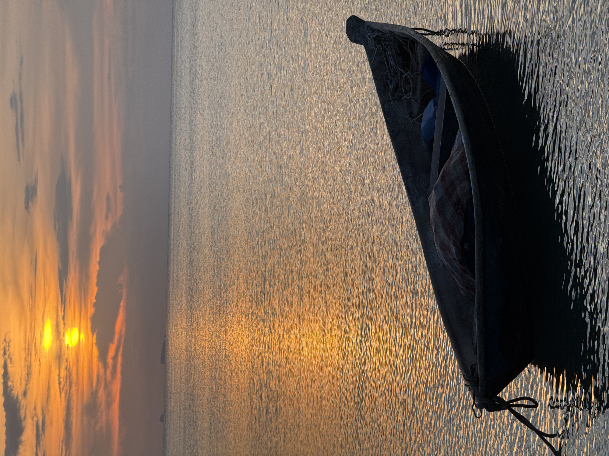A low-angle, vertical shot depicts a tranquil scene at sunset over the water in Ko Samui District, Thailand. The sun, a bright yellow orb, hangs low in the sky, casting a golden, shimmering reflection across the calm sea. Wispy clouds, tinged with orange and pink hues, drift across the horizon. In the foreground, a dark, weathered wooden boat rests on the water, its silhouette stark against the glowing sea. A coiled rope secures the boat, its knot clearly visible. Inside the boat, a checkered fabric is draped over some unseen objects, and blue cushions are visible towards the stern. The water's surface is rippled, creating a textured, reflective surface that mirrors the colors of the sky. Distant, dark shapes that appear to be islands or landmasses are visible on the horizon line. The overall atmosphere is one of peaceful solitude and natural beauty, characteristic of coastal Thailand during twilight.
