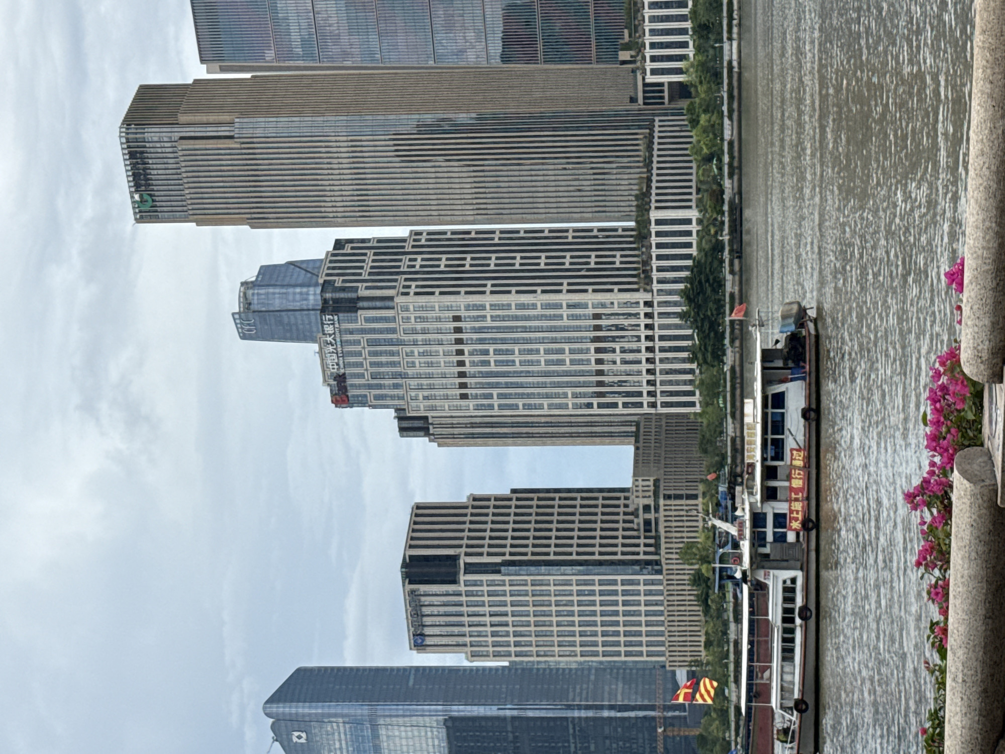 This media file captures a scene of modern architecture and a riverfront setting in Tianhe District, China. The primary subjects are several tall, glass-fronted skyscrapers, characteristic of a contemporary urban landscape. These buildings are characterized by their vertical lines and reflective surfaces, creating a sense of scale and grandeur.
In the foreground, a river flows, with a boat docked at the edge. The boat appears to be a passenger vessel, possibly for tours or transport, indicated by its multiple decks and windows. The river's surface is rippled, suggesting a gentle breeze or current. Along the riverbank, lush green trees add a touch of nature to the urban environment, contrasting with the concrete and glass of the city. A stone railing runs along the right side of the frame, adorned with vibrant pink flowers, adding a splash of color to the scene.
There are no prominent people visible in the frame, suggesting the focus is on the architectural and natural elements of the location. The weather appears to be overcast, with a light, diffused sky, suggesting it could be morning or late afternoon.
Visible text on one of the buildings, translated from Chinese, appears to read "China Everbright Bank," indicating a prominent financial institution. Another sign on the boat reads in Chinese, "水上施工進行," which translates to "Water construction in progress," implying ongoing work or maintenance activity along the waterway. The overall mood is one of urban vitality and development, with a hint of tranquility offered by the river and greenery.