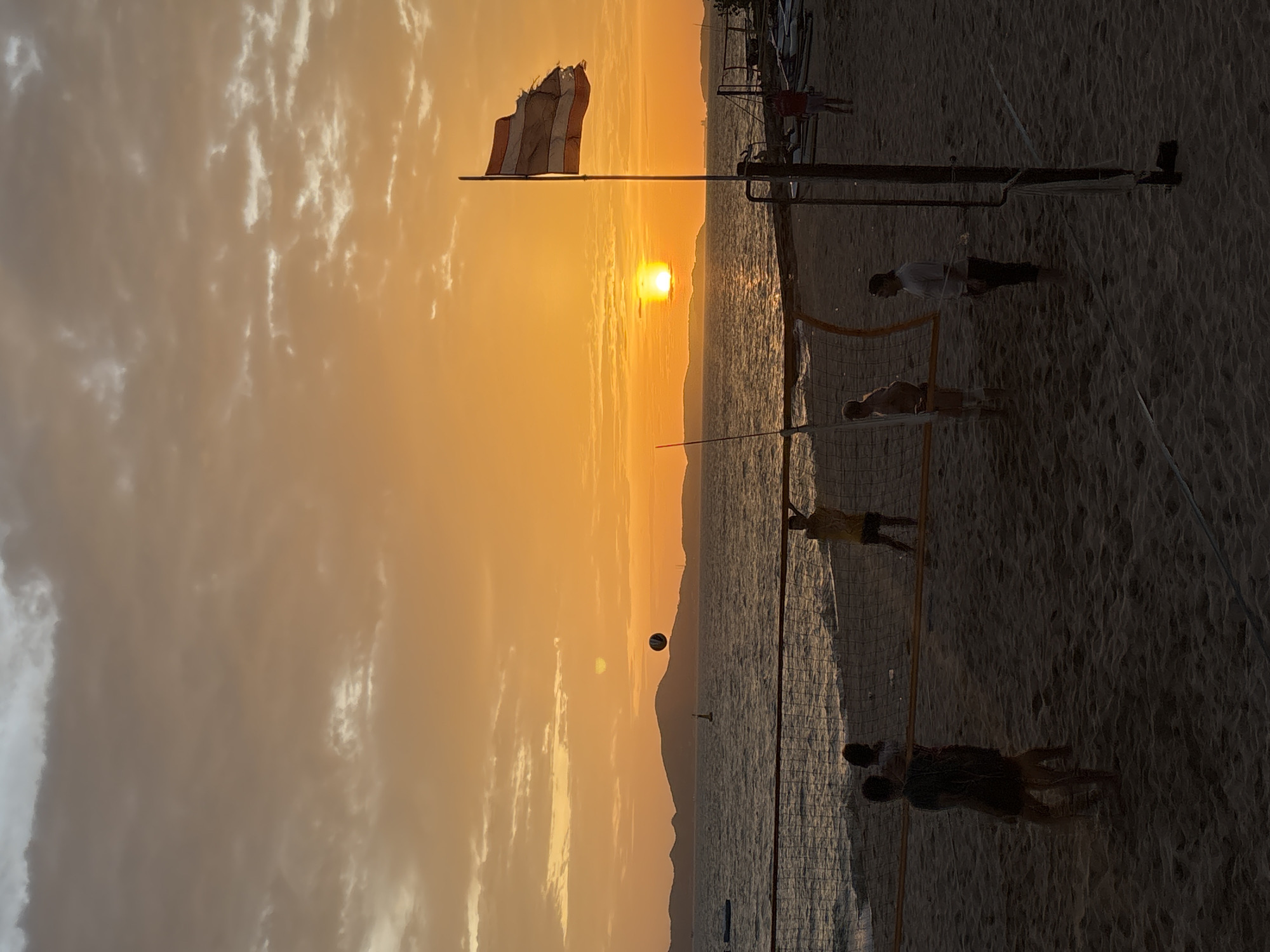 At sunset on a beach in Pattaya City, Thailand, people play volleyball. The sun, a bright orb, is low on the horizon, casting a warm orange glow across the sky, which is filled with scattered clouds. The ocean stretches out before the beach, with gentle waves lapping at the shore. In the foreground, a volleyball net is set up, and several individuals are engaged in a game. A volleyball is in mid-air, suggesting an active play. A Thai flag flutters from a flagpole to the right of the frame. Silhouettes of people and a volleyball net are visible against the bright sky. The scene conveys a sense of leisure and recreation.