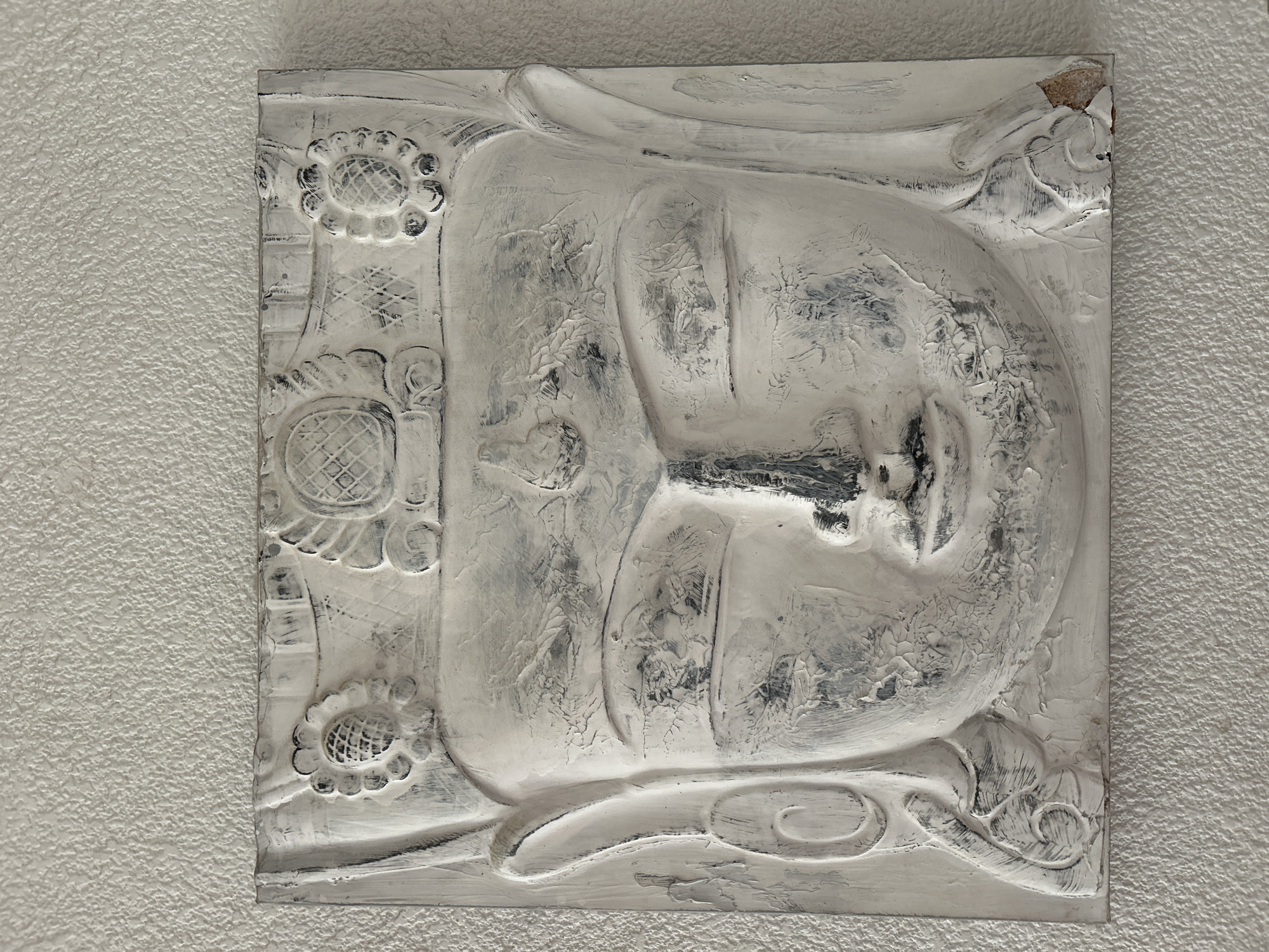 A square-shaped relief sculpture, predominantly white with distressed black and grey undertones, is mounted on a light-colored, textured wall. The central subject of the sculpture is a serene human head, depicted in a three-quarter profile with closed eyes, suggesting sleep or contemplation. Facial features like the nose and lips are discernible.
The head is framed by ornate decorative elements. Along the left side of the sculpture, there are circular, floral motifs, possibly resembling sunflowers, with patterned centers. Swirling, organic patterns adorn the lower and upper right sections of the artwork. The sculpture's surface is heavily textured, showcasing visible brushstrokes and a stucco-like finish, which contributes to its antique or weathered appearance. One corner at the top right of the plaque shows minor damage, revealing a brownish material beneath the white coating.
The overall color palette is monochromatic, relying on shades of white, grey, and black, which enhances its classical aesthetic. No specific activity is taking place, as it is a static art piece. There is no visible text. While the location context provided is Barcelona, Spain, the image itself does not contain any specific visual cues to this city beyond the textured wall, which could be typical of architecture in the region.