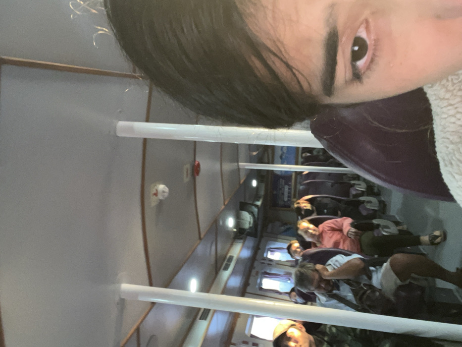 The image captures a selfie taken inside what appears to be a passenger ferry or large boat. The perspective is from a person (likely female, given the eye and dark hair) positioned at the top of the frame, looking down into the vessel's interior. Part of their face, from the forehead to below the eye, is visible, along with dark hair and a light-colored, fluffy collar or hood.

The main scene depicts the interior cabin of the vessel. Rows of dark, upholstered seats extend away from the viewer, occupied by several passengers. The aisle runs down the middle, separating the seats. The passengers are casually dressed; some appear to be looking down or resting, while others look forward. Visible individuals include a person in a salmon-colored shirt, another in a white shirt, and one wearing a cap.

The cabin features light-colored walls, possibly light grey or beige, with wooden trim. White horizontal poles are visible overhead, likely part of the ceiling structure or handrails. Recessed ceiling lights illuminate the space. On one wall, a red fire alarm pull station and a white smoke detector are clearly visible, indicating safety provisions. Further down the cabin, rectangular windows allow ambient daylight to filter in.

The activity taking place is travel by water, with passengers seated during their journey. The overall mood is calm and routine. Given the context of Hà Tiên, Vietnam, the setting is likely a ferry traversing coastal waters or rivers. No distinct text is legible within the image.