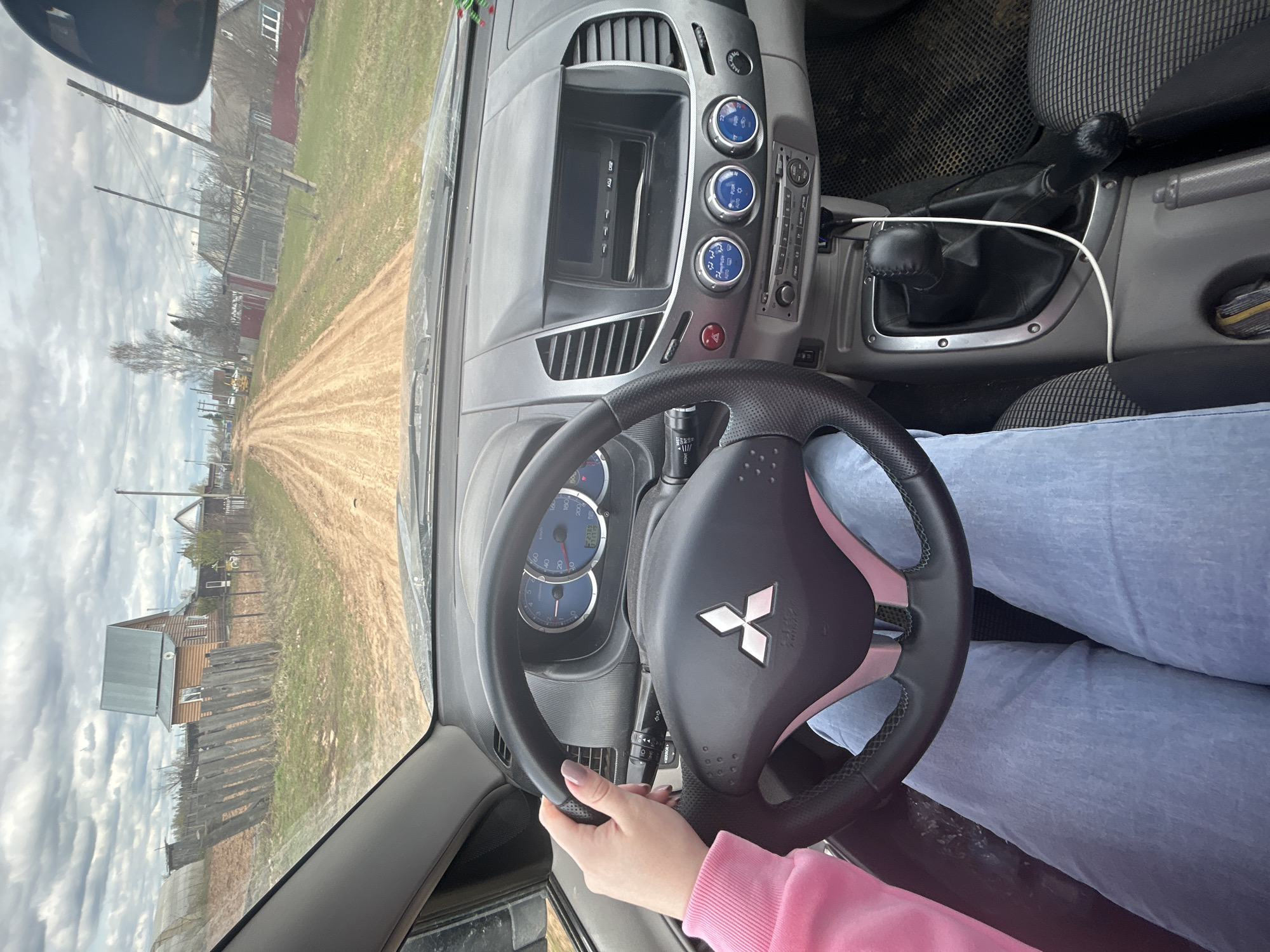 The image shows the interior of a Mitsubishi L200 car, with the steering wheel in focus. A person with light blue jeans and a pink sweater is holding the steering wheel. The car is driving down a dirt road in Фоки, Russia. Wooden houses and fences line the road. The sky is cloudy.