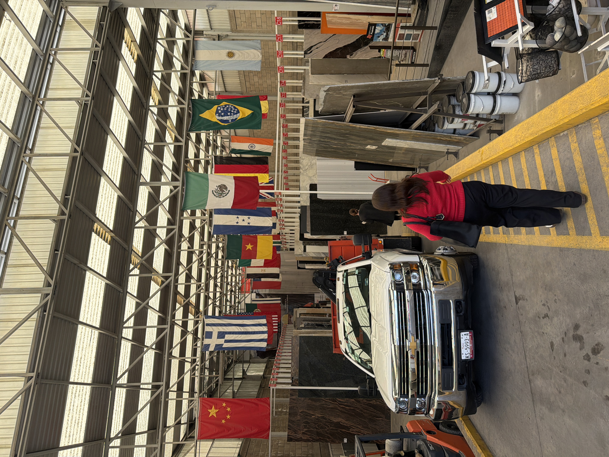 The image shows the interior of a warehouse in Puebla, Mexico, decorated with various international flags hanging from the ceiling. A white pickup truck is parked inside the warehouse, and a woman in a red sweater and black pants is standing near it. Pallets of stone slabs are stacked nearby. The building has a metal roof structure with numerous skylights providing natural light. A forklift is partially visible in the foreground.