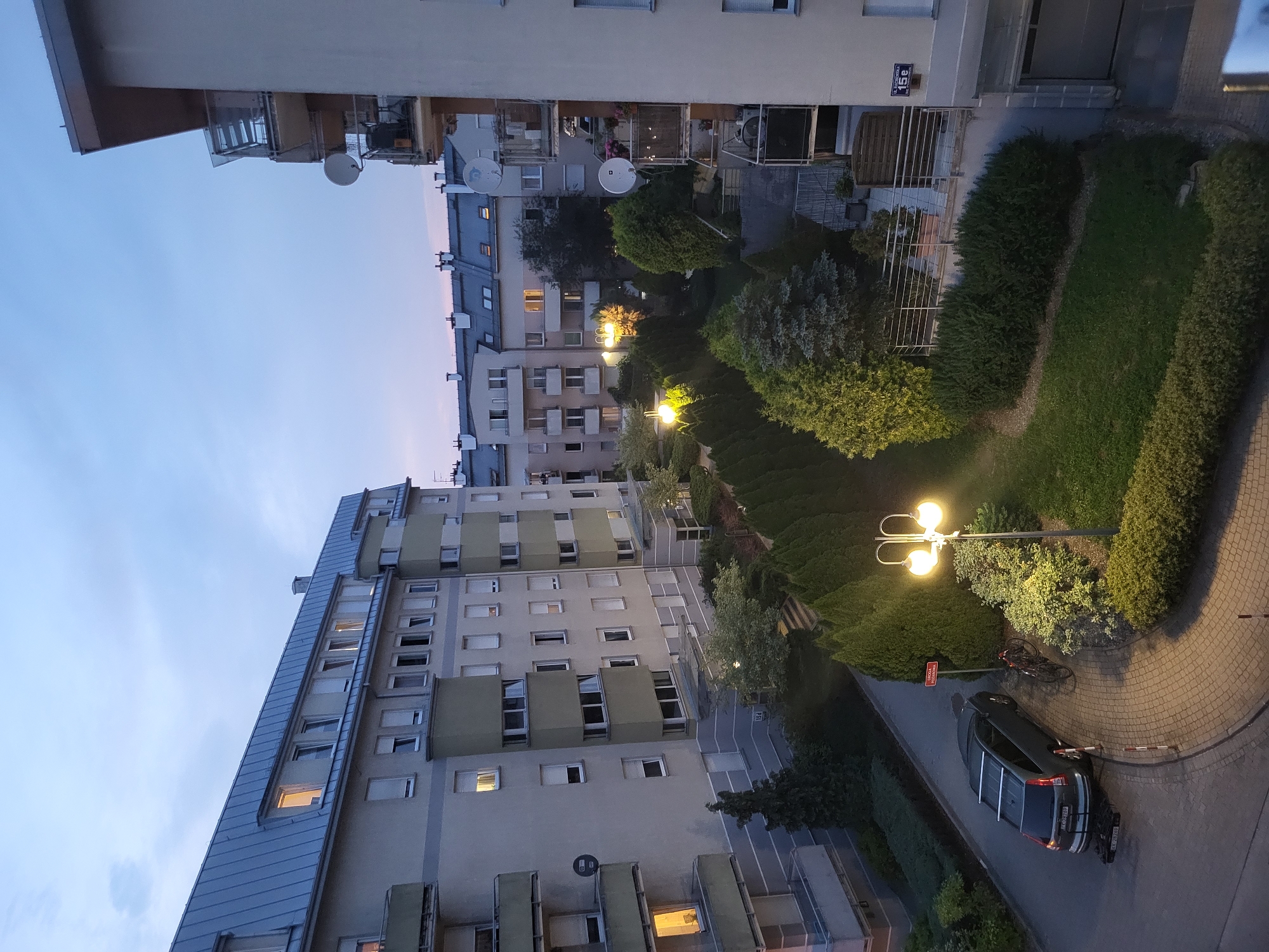 The image depicts a courtyard between apartment buildings in an urban setting at dusk. A dark grey station wagon is parked on a cobblestone driveway, with a bicycle leaning against a street lamp post. The courtyard is landscaped with manicured evergreen trees and bushes. Several apartment buildings with balconies and satellite dishes are visible, with some windows illuminated. The sky is a pale blue with hints of pink and orange from the setting sun. A street sign with Polish lettering is visible on the right side of the image. The scene appears calm and residential.