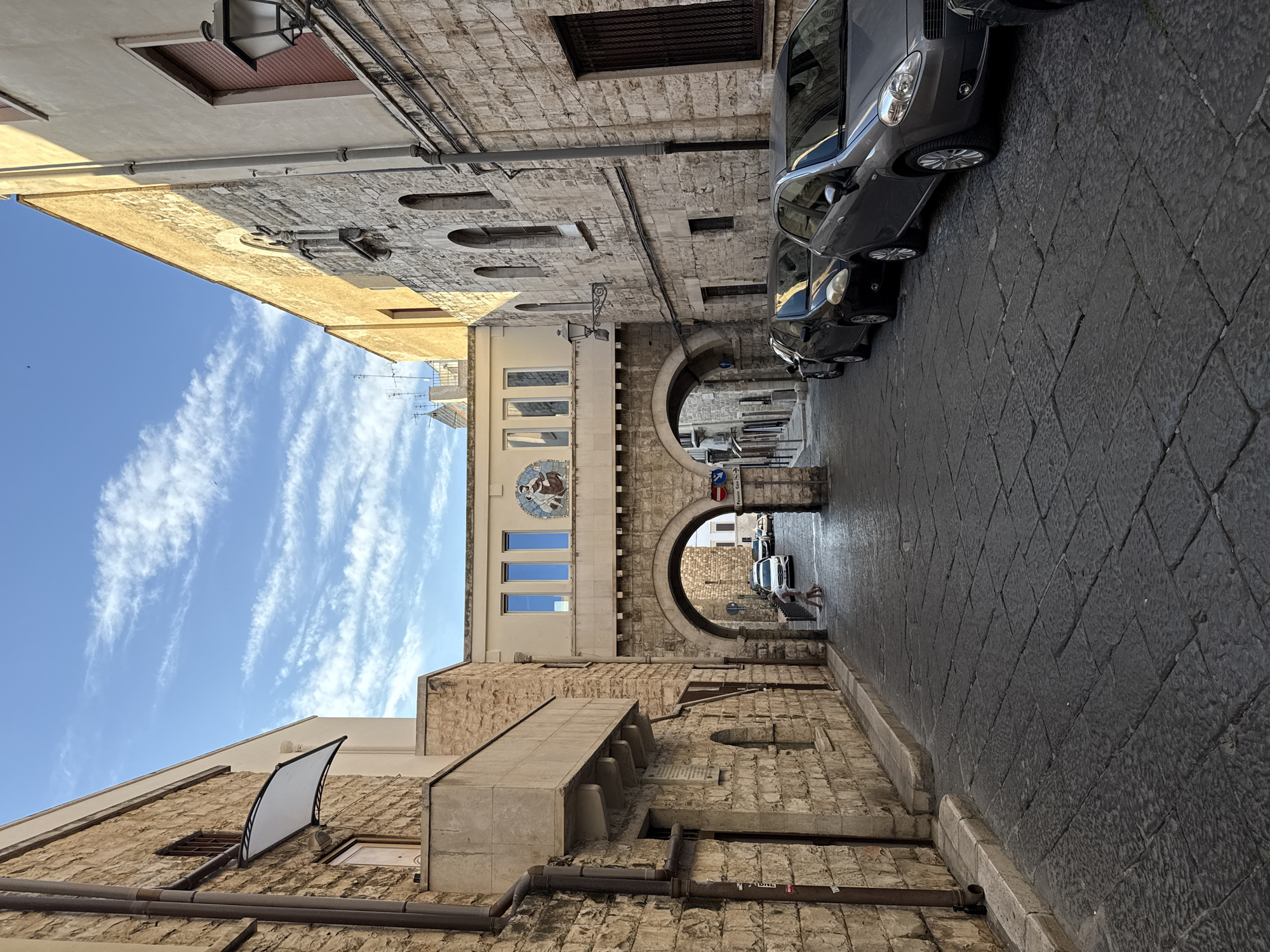 The media file captures a view of a narrow, historic street or alley lined with old stone buildings, under a clear blue sky with wispy white clouds. The image is taken during daylight hours, suggesting good weather conditions.
On the right side of the street, several dark-colored compact cars are parked closely together on a cobbled surface. The buildings are primarily constructed from rough-hewn, light-colored stone blocks, with visible pipes and conduits running along their exteriors.
A prominent feature of the scene is a series of archways that lead further down the street, creating a tunnel-like passage. Above the first archway, on a smoother, lighter-colored building section, there is a circular mosaic or mural depicting two figures. This building also features three rectangular windows below the mosaic.
Beyond the first archway, a second, larger archway is visible. A single person is sitting in the shaded area on the right, under this second archway. A blue directional sign is mounted just past the first archway on the right, displaying "Piazza Cavour" with a right-pointing arrow. Various windows, some with grates, are seen on the building facades, and a modern-looking light fixture is attached to an upper section of a building on the left. The street itself is paved with irregularly shaped stones.