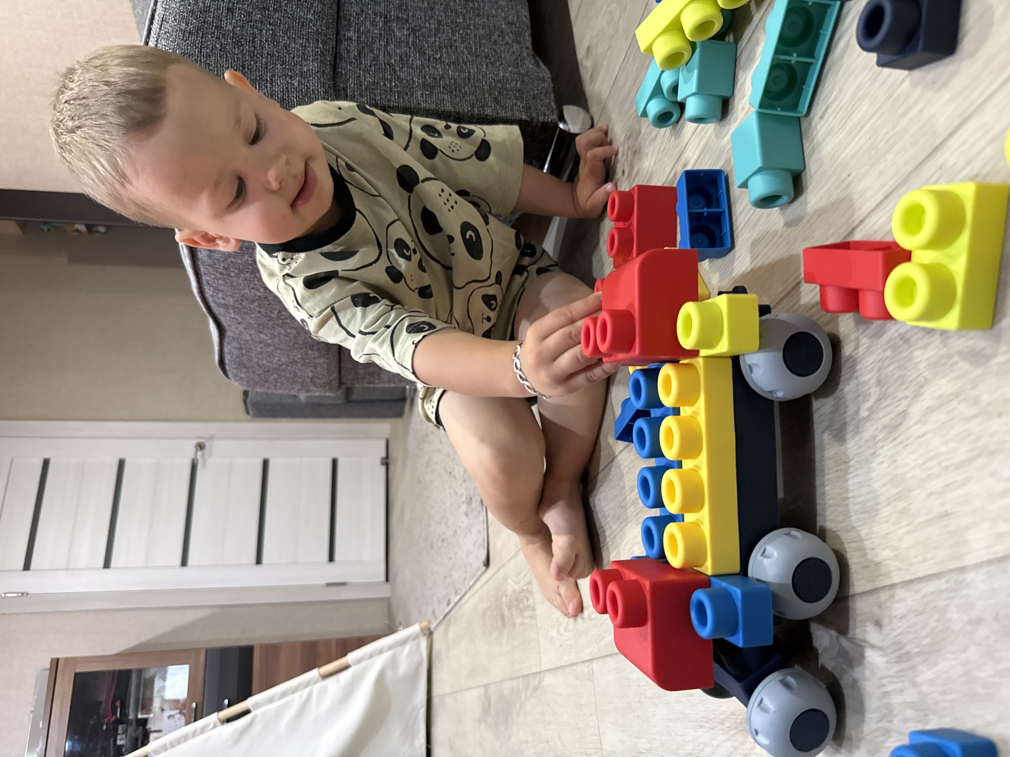 In Rusanovo, Russia, a young Caucasian boy with blond hair sits on a light wood floor playing with colorful building blocks. He is wearing a light-colored t-shirt with a pattern of black panda faces. In front of him is a partially assembled toy train made of red, yellow, blue, and dark gray blocks, with gray wheels. Scattered around the boy are additional building blocks in various colors including green, blue, and yellow. The background shows a portion of a gray sofa, a white interior door, and a wooden cabinet. A white teepee-like structure is partially visible to the left of the frame. The lighting suggests an indoor setting, possibly a living room or playroom.