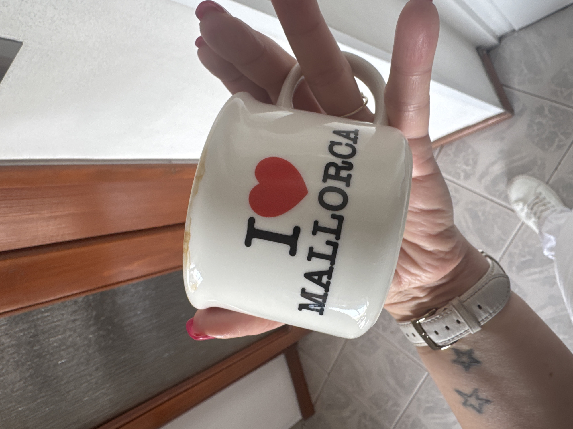 A person holds a white mug with the text "I ❤️ MALLORCA" vertically printed on it. The mug is held in front of a background consisting of a wooden surface and a tiled floor. The person wears a white watch and has two star tattoos on their forearm. The image provides no information about the city of Sofia, Bulgaria.