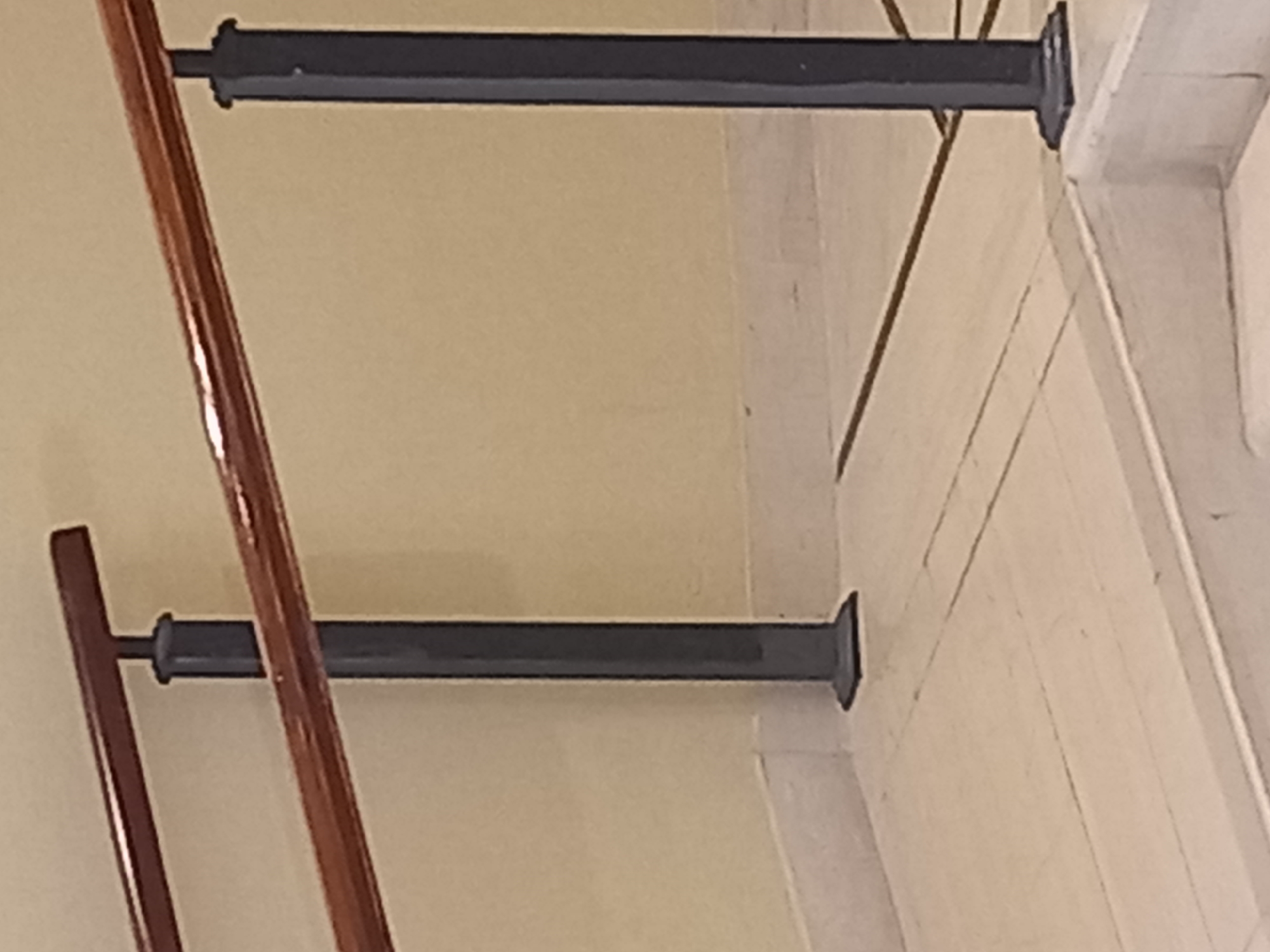 The image shows a close-up view of a dark gray metal handrail with a wooden top, supported by dark gray vertical posts. The handrail is installed on a beige wall and is positioned over a light-colored tiled floor. There are two visible posts supporting the handrail. The floor has some dark lines suggesting grout or inlay patterns. In the foreground, the top of white marble stairs is visible. The scene appears to be indoors, possibly in a public building or staircase. The image does not depict any people or provide specific indicators of the location within the city of Kos, Greece.