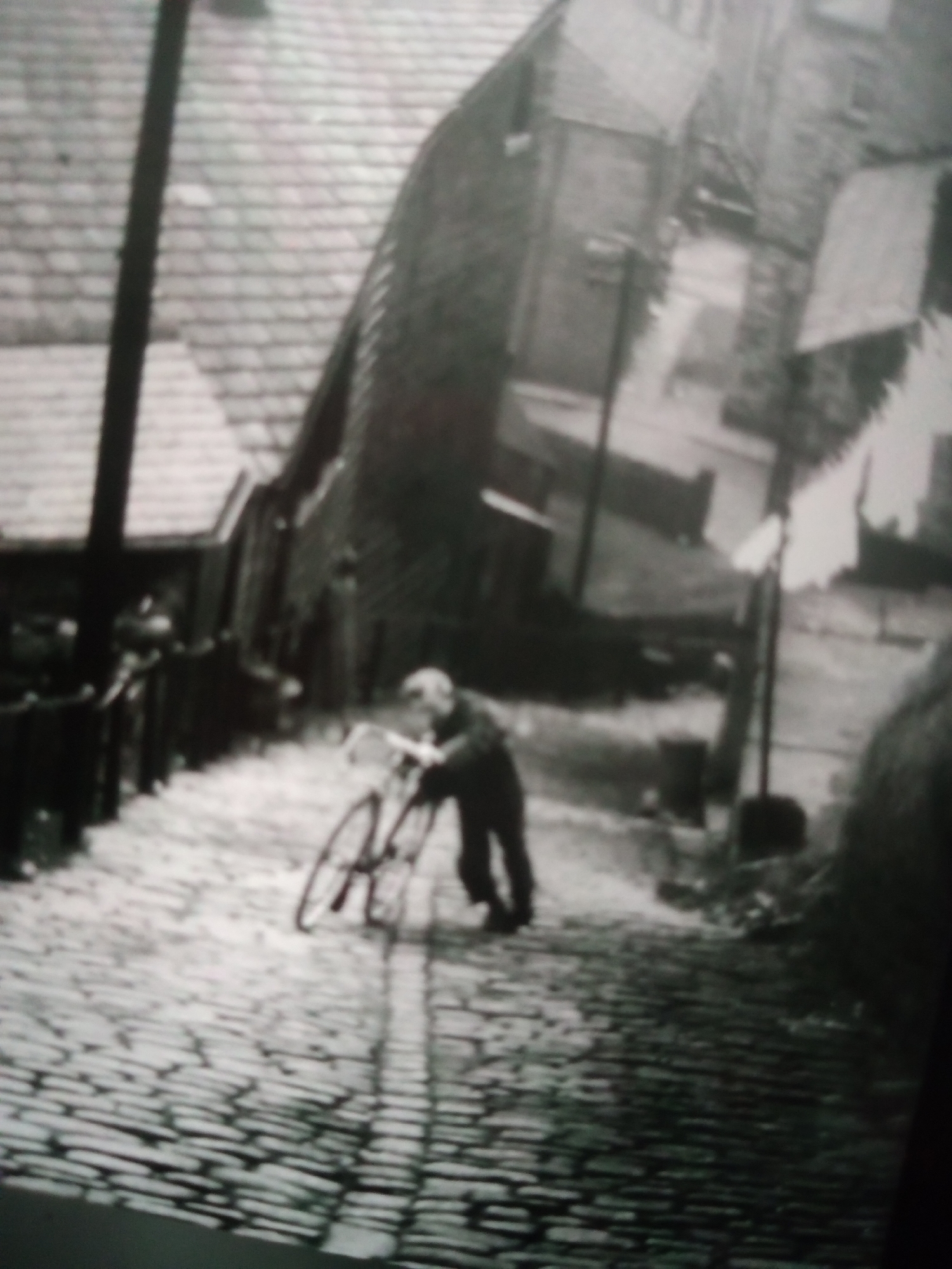 A black and white, low-resolution, slightly blurry photograph depicts a cobblestone street with a person pushing a bicycle uphill. The person, appearing to be an adult male based on the silhouette and clothing, is bent over the handlebars. They are wearing dark clothing and possibly a cap or hat. The bicycle is upright and its wheels are visible. The street is paved with cobblestones, indicating an older urban setting. Buildings with visible roofing and windows line the street, suggesting a residential or commercial area. Laundry hangs on a line in the background, adding to the sense of daily life. A utility pole is also present on the left side of the frame. The overall impression is of a historical street scene.