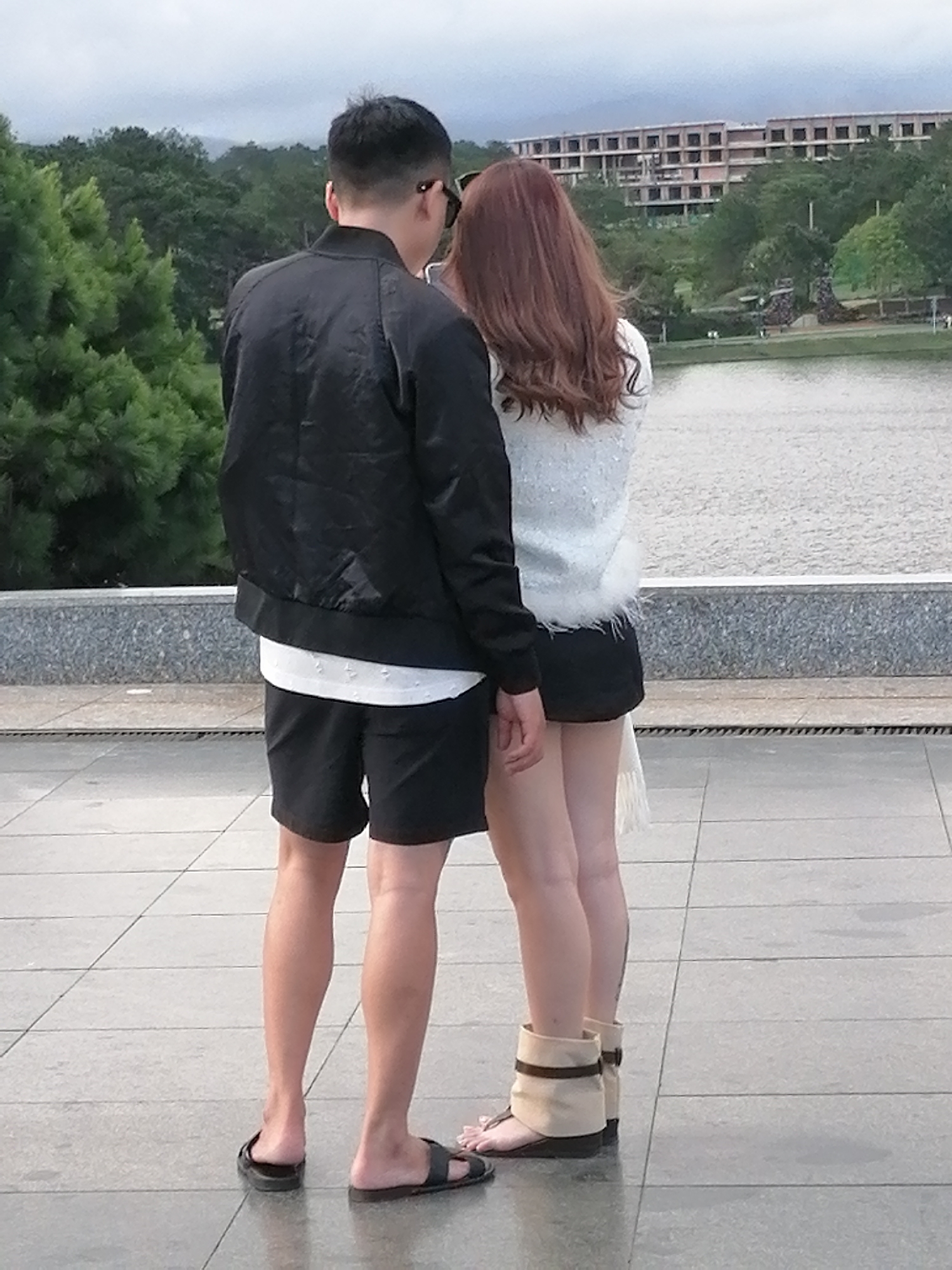 A man and a woman are standing back-to-back on a tiled patio, possibly at Xuan Huong Lake in Da Lat, Vietnam. The man is wearing a black bomber jacket, a white t-shirt, and black shorts. He has short, dark hair and is wearing black sunglasses. The woman has long, auburn hair and is wearing a light-colored, fuzzy sweater over a black skirt. She is wearing tan ankle boots with dark straps. A large body of water, likely Xuan Huong Lake, is visible behind them, with trees and a multi-story building under construction on the far bank. The sky is overcast.