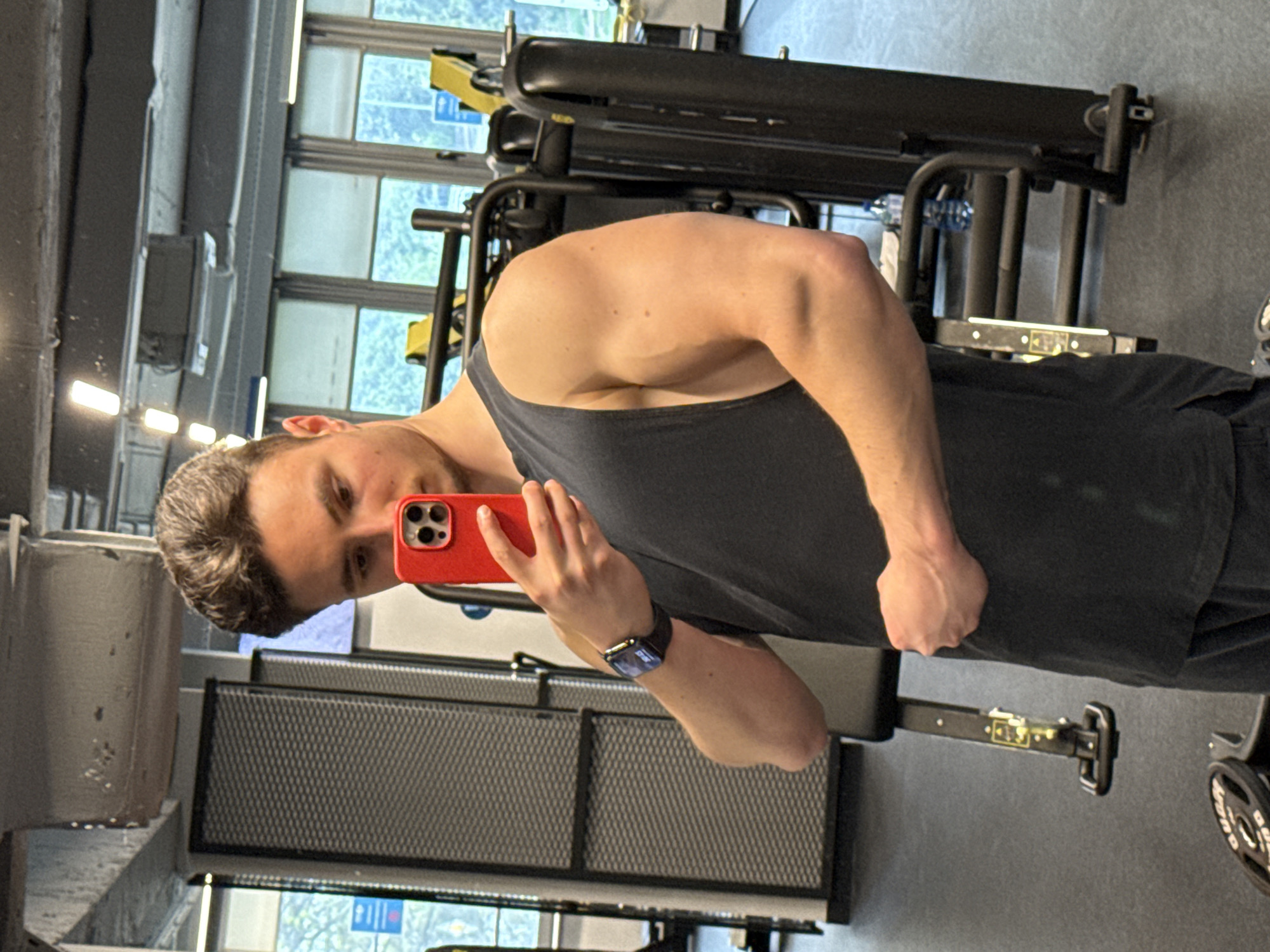 A man is taking a selfie in a gym. He is wearing a dark tank top and shorts and has a smartwatch on his left wrist. He is holding a red iPhone in his right hand. He is standing in front of gym equipment, including weight machines and a bench. The background shows windows with a view of the outside. The location is likely in Sofia, Bulgaria.