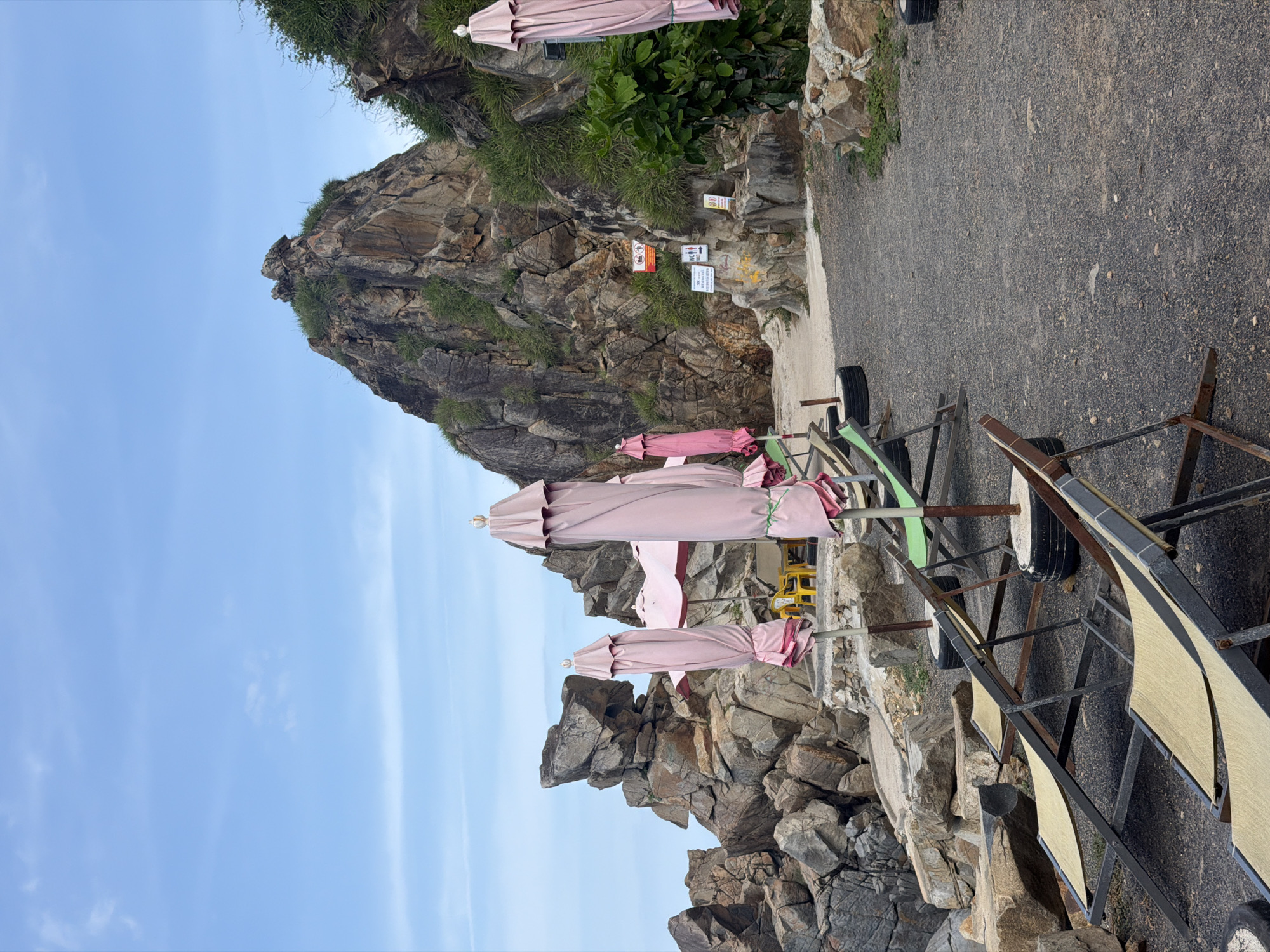 The image depicts a rocky coastline with several beach chairs and closed pink umbrellas arranged on a gravel path. In the background, large rock formations rise, with some vegetation growing on them. A sign is visible on the rock face, indicating "WC" and "TOILETS". The sky is cloudy with a light blue hue. The scene appears to be a quiet, possibly off-season, beach area in Vĩnh Lương, Vietnam, with amenities for beachgoers, but currently unoccupied.