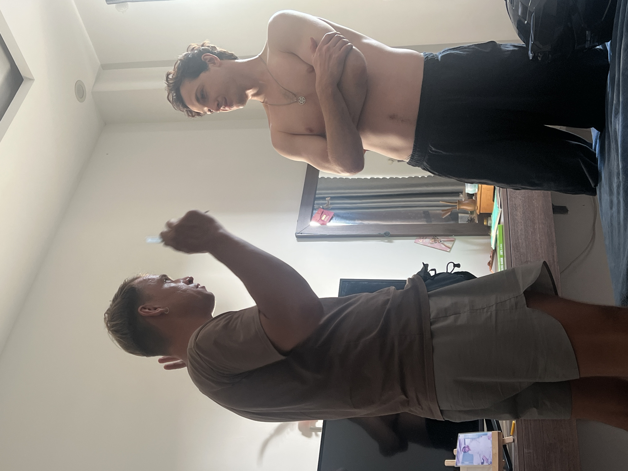 In Đà Nẵng, Vietnam, two men are in a brightly lit room. The man on the left, wearing a brown t-shirt and grey shorts, stands with his left arm raised and his right hand holding an object, possibly a pen or stylus, while gesturing with his left hand. He has short, light-colored hair and an earring. The man on the right, shirtless and wearing black shorts, stands with his arms crossed and is looking towards the left. He has curly brown hair and a necklace. Behind them, a wooden desk is visible with various items including a television, a mirror, and some stationery. The environment appears to be a bedroom or living space.