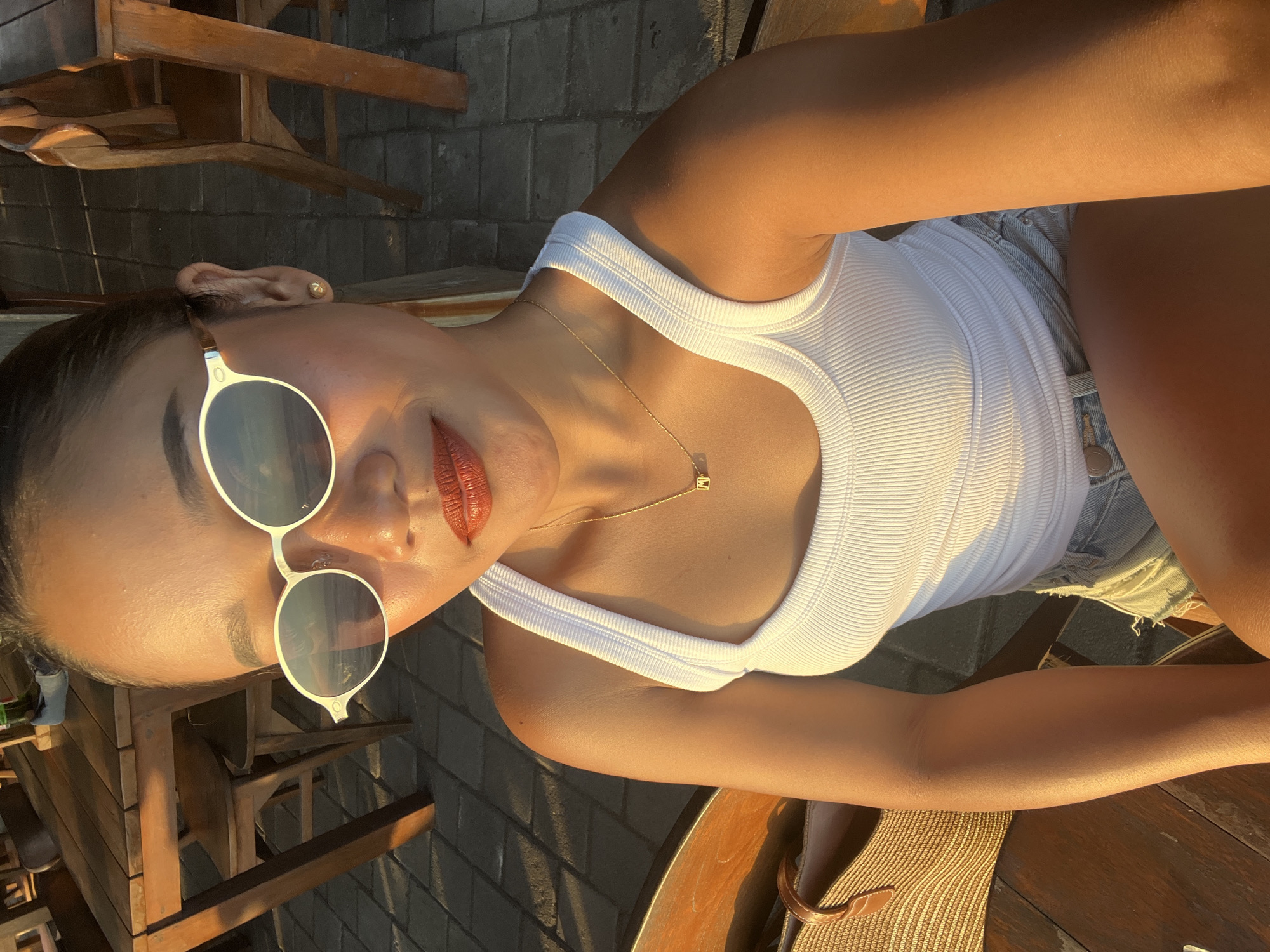 A woman is pictured from the chest up, sitting at a wooden table outdoors. She is wearing a white tank top, denim shorts, and white-rimmed sunglasses. A gold necklace with a pendant is visible around her neck. The setting appears to be a beachside restaurant in North Kuta, Indonesia, with wooden chairs and tables visible in the background. The sun is low, casting a warm glow on the scene.