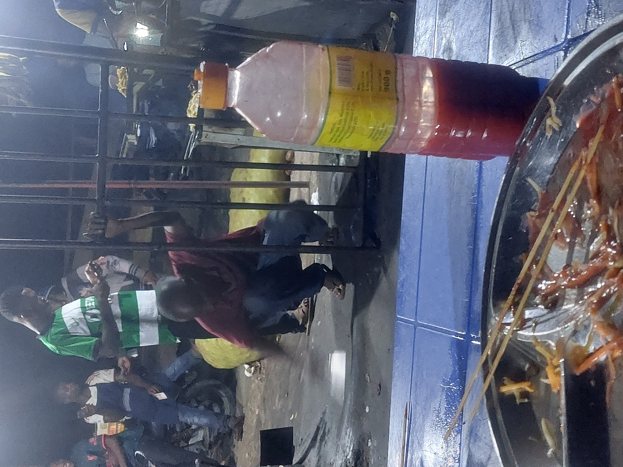 This is a nighttime street scene in Dar es-Salaam, Tanzania. The focus includes a metal cage behind which people mill around. In the cage, a man in a red shirt and dark pants bends over. In front of the cage, on a blue plastic table, there is a large bottle filled with a two-toned liquid (pink and red). To the right of the bottle sits a black pan filled with cooked food on skewers. Other people are visible in the background, including a man in a green and white striped shirt. In the foreground are other people, some on motorcycles. There are street lights visible, illuminating the scene.