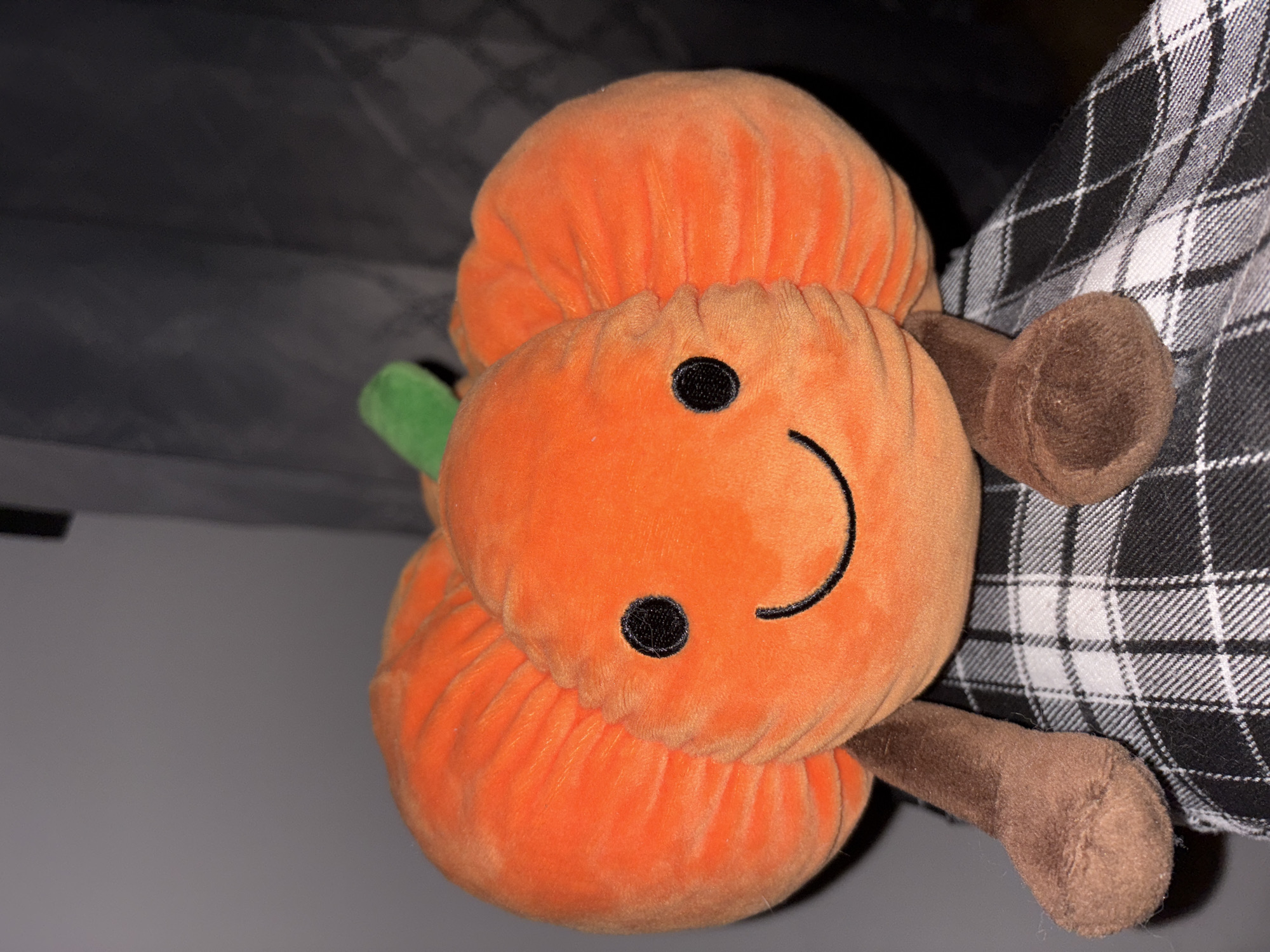 A plush toy resembling a stack of pumpkins is positioned in front of a black and white plaid fabric. The toy features two orange, rounded sections stacked atop each other, with a green stem extending from the top. The middle section of the toy has two black button eyes and a curved, stitched smile. Two brown plush arms extend from either side of the middle pumpkin. The background consists of a light gray wall on the left and a darker gray fabric or surface above. The location is not evident from the image.