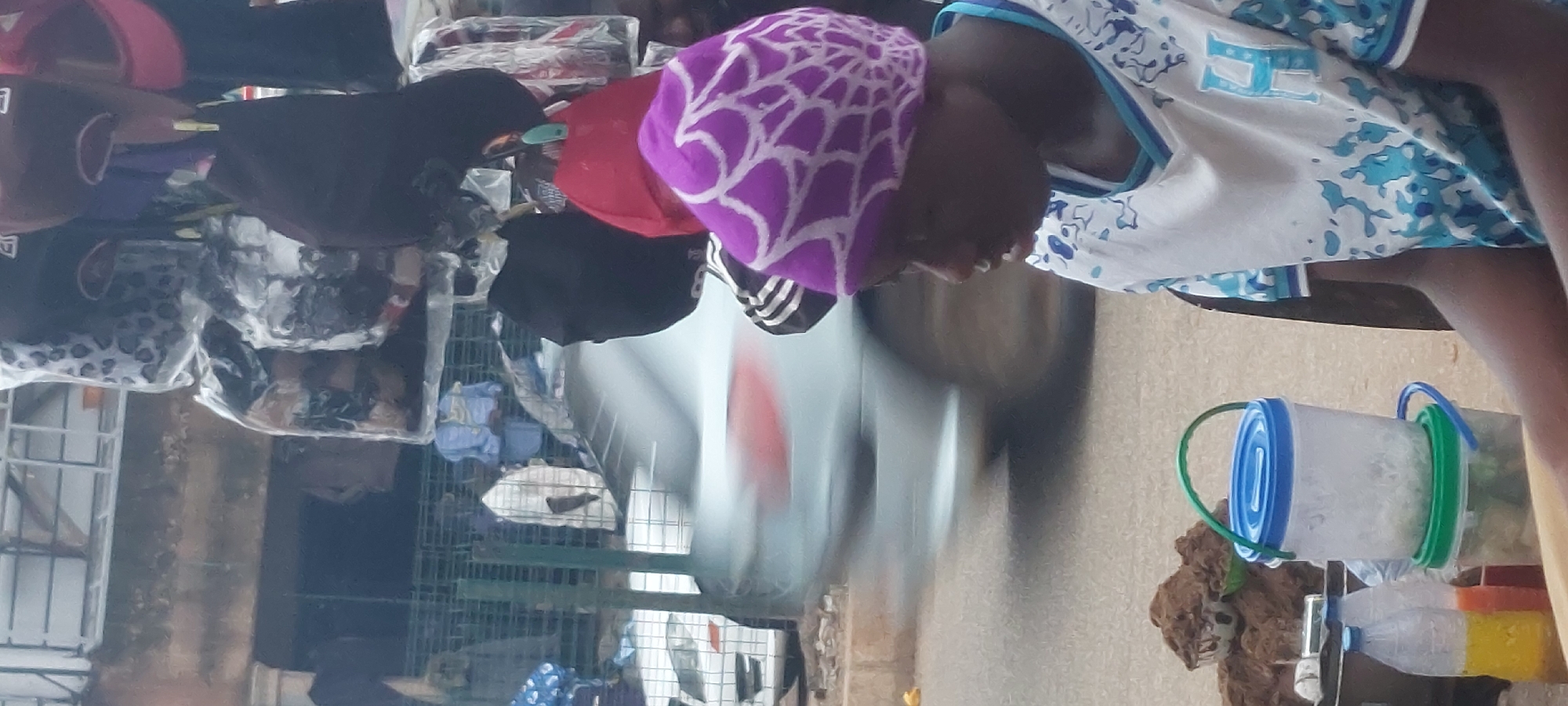 The image depicts a street scene in an unknown city. In the foreground, a person wearing a purple headscarf with a white web-like pattern and a white t-shirt with blue designs is visible from the side. The person appears to be seated, with their arms resting on a surface. To the left of the person, there are several plastic containers. One is a clear container with a blue lid, filled with what appears to be shredded material, stacked on top of another clear container with a green lid, which also contains shredded material. Two plastic bottles, one with yellow liquid and the other with orange liquid, are placed in front of the containers. In the background, a silver car is blurred due to motion, suggesting it is driving past. Behind the car, there is a stall or vendor display with various hats hanging. Some hats are covered in plastic bags, while others are exposed. The overall environment appears to be an outdoor market or roadside setting.