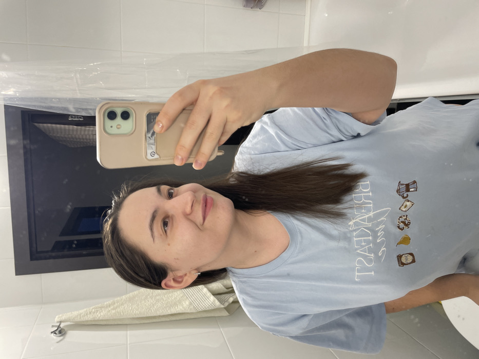A single female is captured in a mirror selfie within a domestic bathroom in what is understood to be Соболиха, Russia. The subject, with dark brown hair, wears a light blue t-shirt with reversed text "BREAKFAST time" and various breakfast-themed icons (jar, pear, pastries, coffee pot) printed on the front. She holds a light tan-colored smartphone with a light green camera module in her right hand, positioned to capture her reflection. Her left ear displays a small hoop earring. She presents a faint smile, looking slightly to her right, away from the camera lens. The bathroom features white tiled walls, a silver towel hook with a light-colored towel draped over it to the left, and a dark brown doorframe visible in the mirror's reflection. A clear shower curtain and a white bathtub are also partially reflected on the right side of the image.