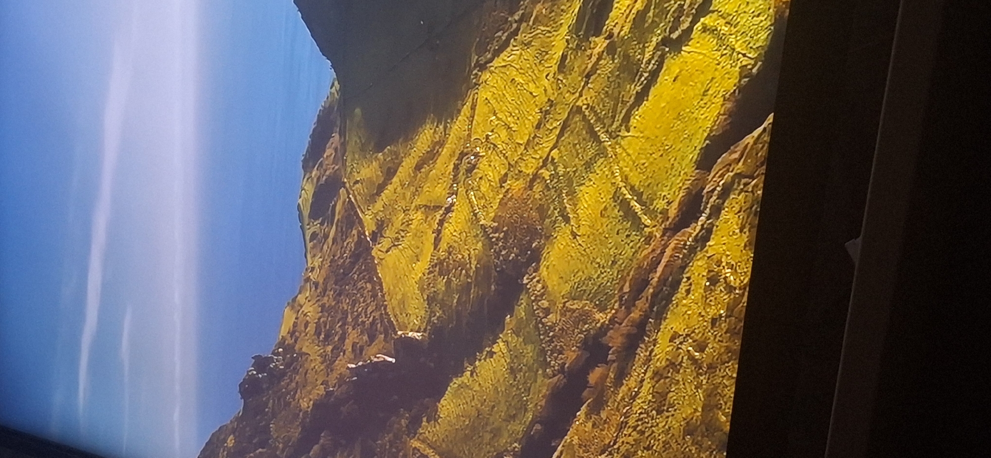 The image shows a sweeping, high-angle view of a vibrant green, terraced landscape under a bright blue sky with thin white clouds. The terrain is characterized by rolling hills that have been meticulously carved into a series of distinct agricultural fields, divided by stone walls or natural contours. The sunlight highlights the rich, golden-green hue of the vegetation, suggesting a pastoral or agricultural setting. In the distance, a vast expanse of deep blue water meets the horizon, possibly an ocean or a large lake. The overall impression is one of natural beauty and human cultivation. There are no discernible people, animals, or specific structures in this image, making it difficult to pinpoint an exact location. It is not possible to identify any specific landmarks or features that would definitively place this scene within the city of Bila Tserkva, Ukraine. The provided image does not contain any elements that can be attributed to Bila Tserkva.