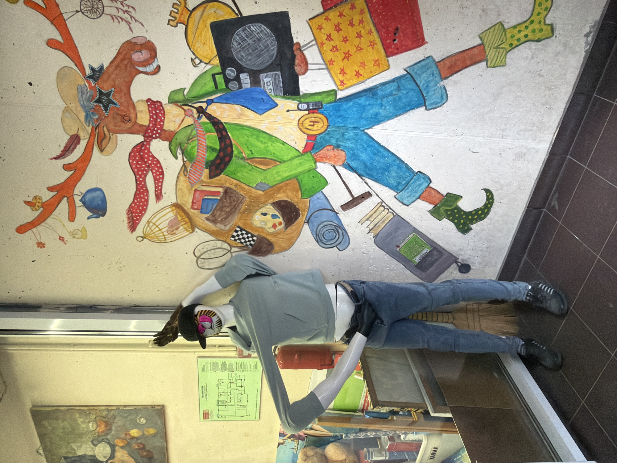 The image shows an indoor scene with a mannequin standing near a wall mural. The mannequin is wearing jeans, a gray shirt, and a black hat. It is positioned next to what appears to be a shelf or display case filled with various items, including books and containers. The wall mural features a whimsical depiction of a character with elements of a moose or reindeer, adorned with various objects like a dreamcatcher, birdcage, and luggage. The mural is colorful and cartoonish, adding a playful element to the space. Based on image recognition and available information, the location is not identifiable as the city of Moscow, Russia.