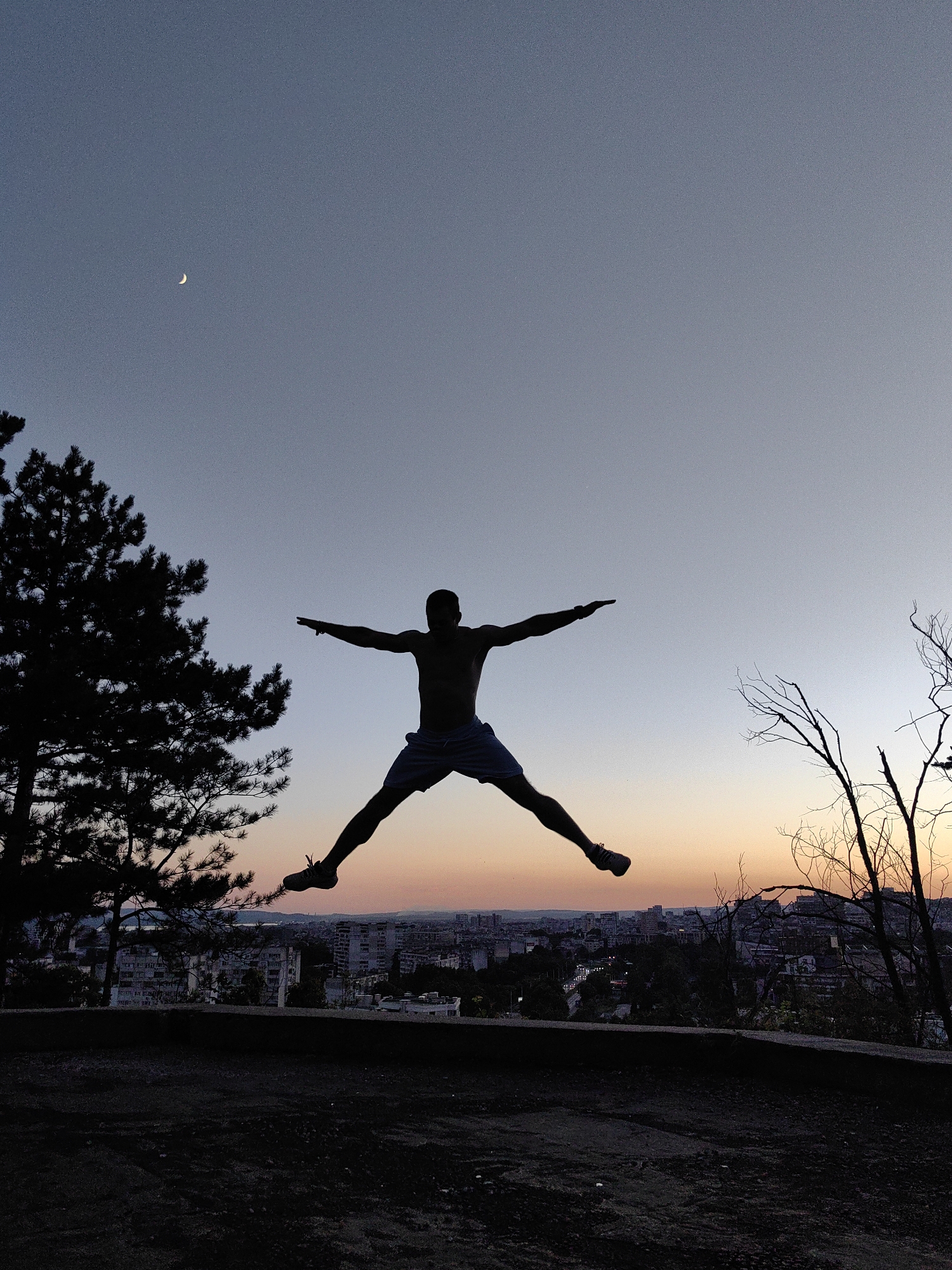 A silhouette of a shirtless person in mid-air jump, legs spread wide and arms extended horizontally, is captured against a dusky sky. The person is wearing shorts. The background features a crescent moon in the upper left quadrant of the sky, a dark silhouette of a pine tree to the left, and the cityscape of Varna, Bulgaria, with buildings and distant hills visible. Bare branches of a tree are visible on the right. The foreground is dark and appears to be uneven ground, possibly a hill or vantage point. The lighting suggests either dawn or dusk, with a warm glow on the horizon.