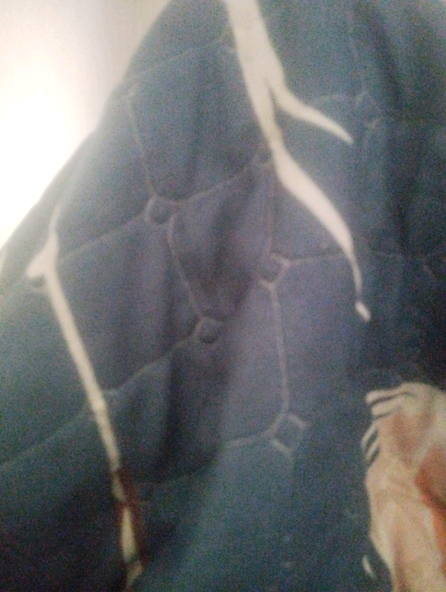 The image depicts a close-up, blurry view of a dark blue, quilted fabric. The quilting creates a pattern of interconnected, rounded hexagonal shapes. A thin, off-white line runs diagonally across the fabric, appearing to be part of a seam or a decorative element. In the bottom right corner, a blurred section of what appears to be skin or a light-colored fabric with texture is visible. The lighting suggests an indoor setting, with a bright light source in the upper left portion of the image. There is no discernible information about the city of Fergana, Uzbekistan, within the image itself.